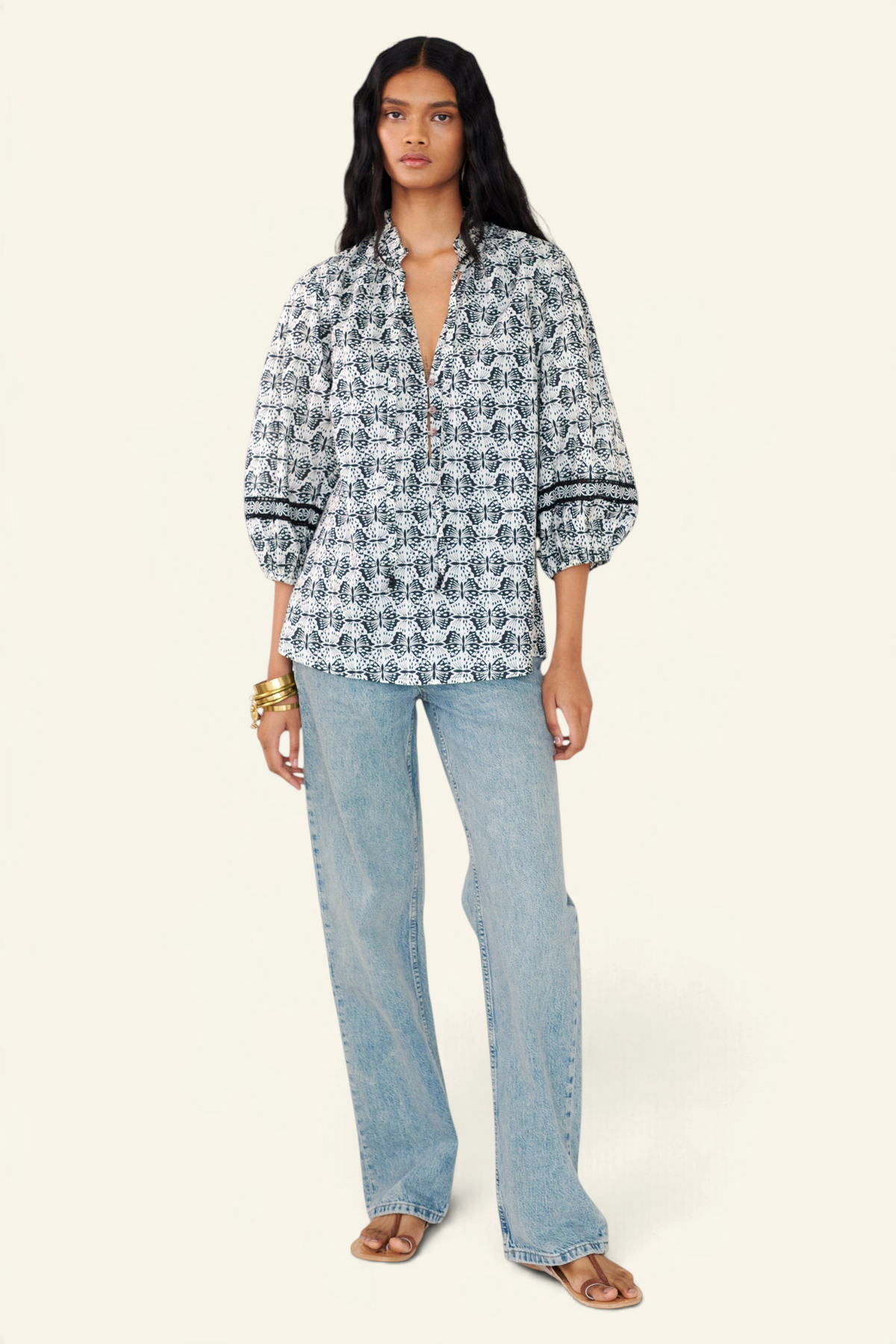 Front view of a woman standing wearing the Lorna top in Flutter print, featuring a ruffled V-neck with adjustable tie, Celtic-knot lace sleeve inserts, and a loose cotton voile silhouette styled with jeans