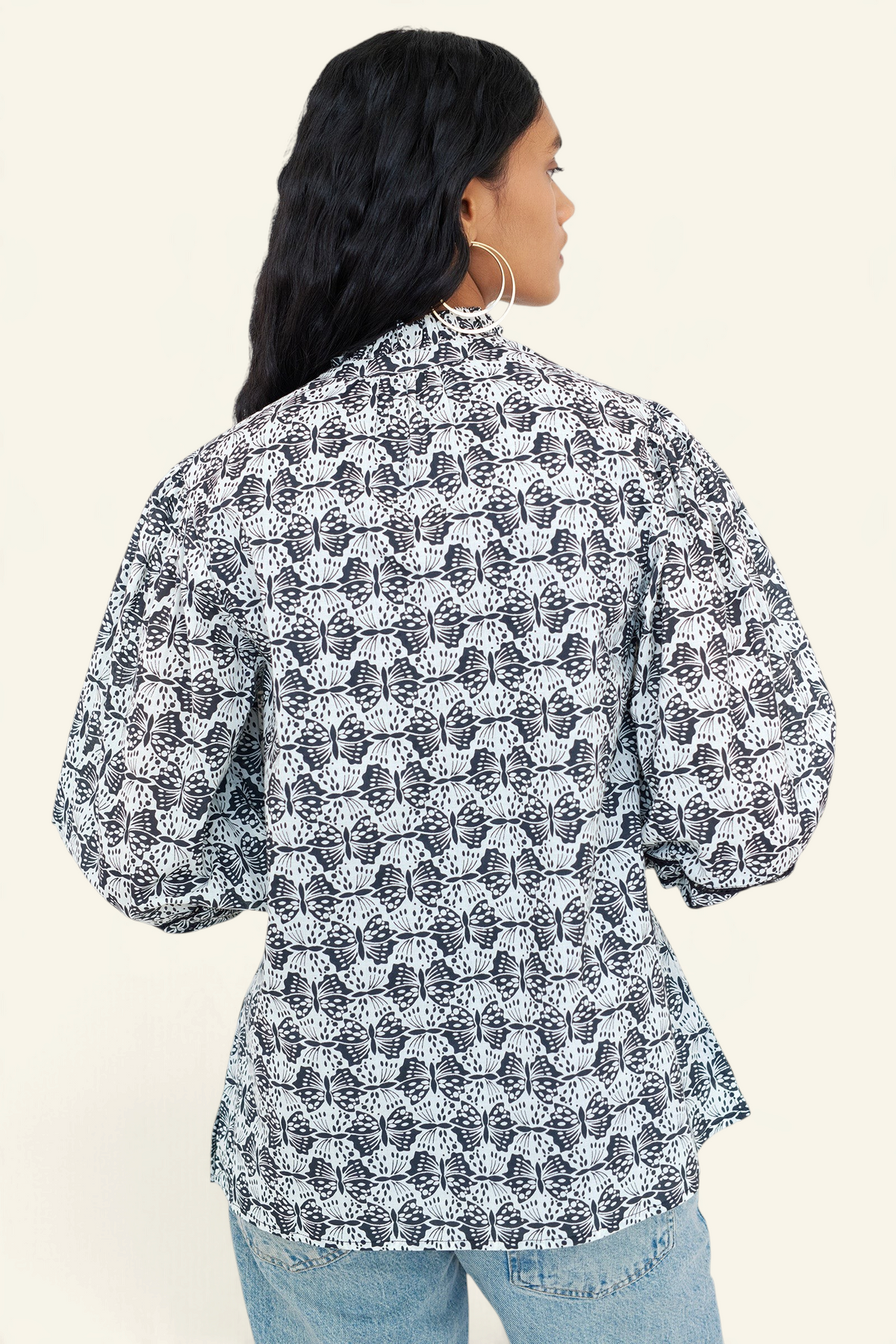 Back view of a woman standing wearing the Lorna top in Flutter print, featuring a ruffled V-neck with adjustable tie, Celtic-knot lace sleeve inserts, and a loose cotton voile silhouette styled with jeans