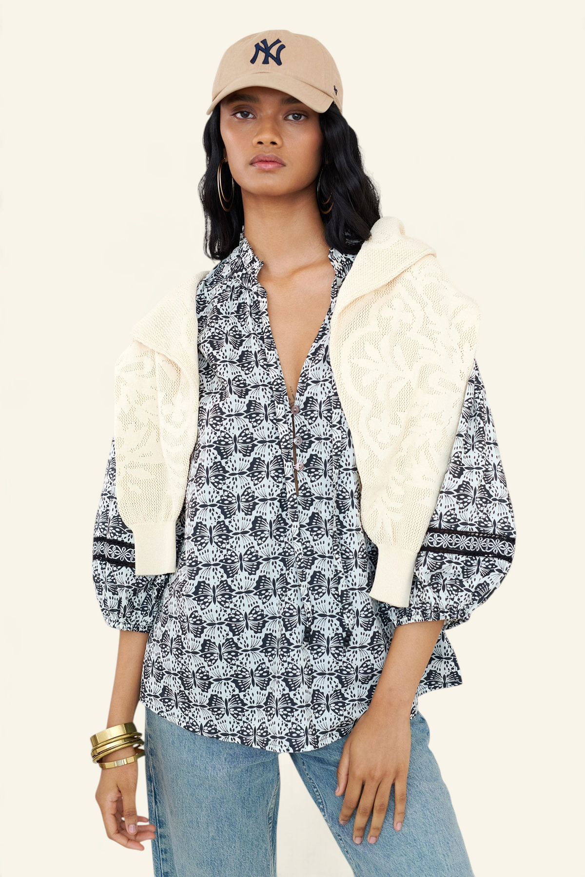 Front view of a woman standing wearing the Lorna top in Flutter print, featuring a ruffled V-neck with adjustable tie, Celtic-knot lace sleeve inserts, and a loose cotton voile silhouette styled with jeans, cap, layered with a sweater on shoulders.