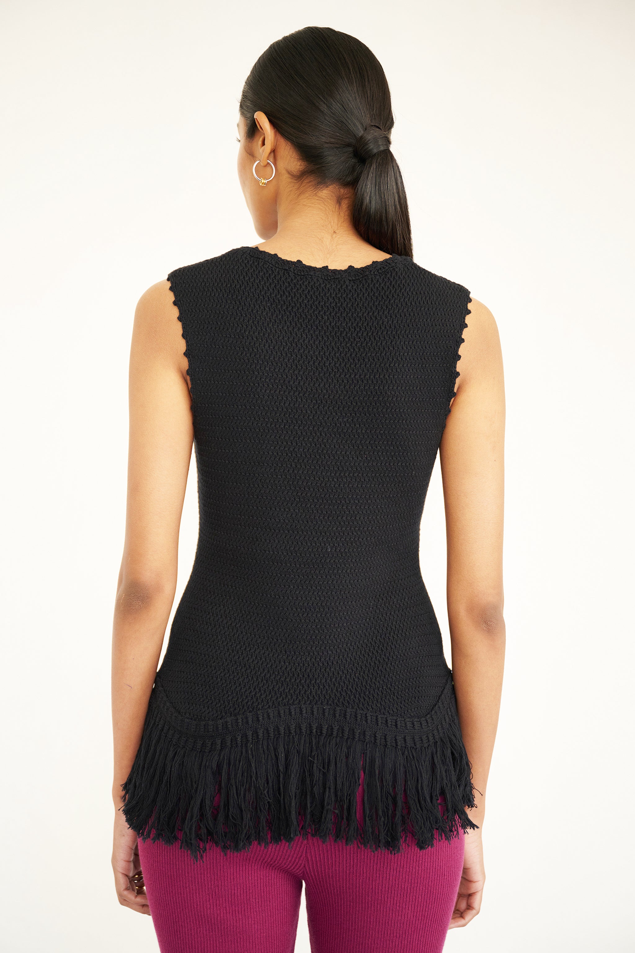 Back view close up of a woman standing. She is wearing Luzia Fringe Tank. It is a black sleeveless cotton knit tank with hand-crocheted trim and a fringed hem, styled for a textured, artisanal look.