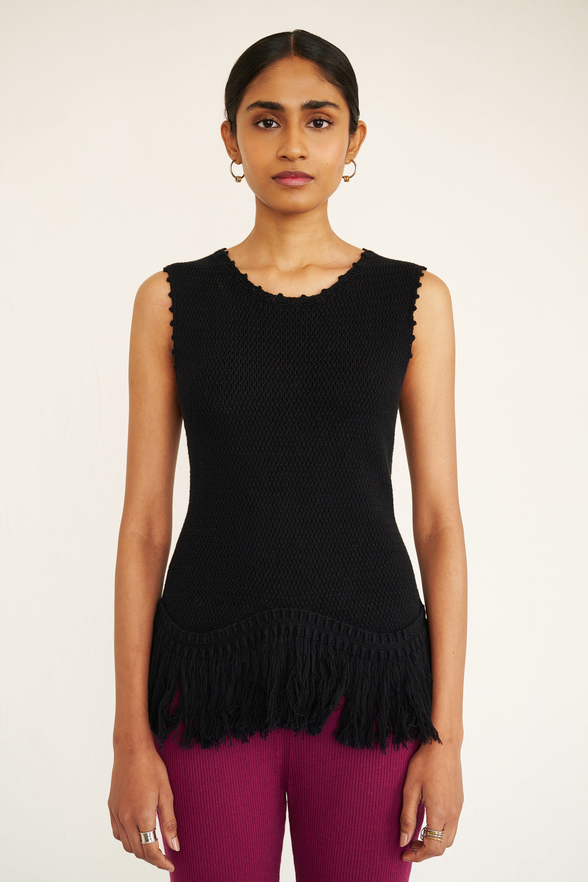Front view close up of a woman standing. She is wearing Luzia Fringe Tank. It is a black sleeveless cotton knit tank with hand-crocheted trim and a fringed hem, styled for a textured, artisanal look.