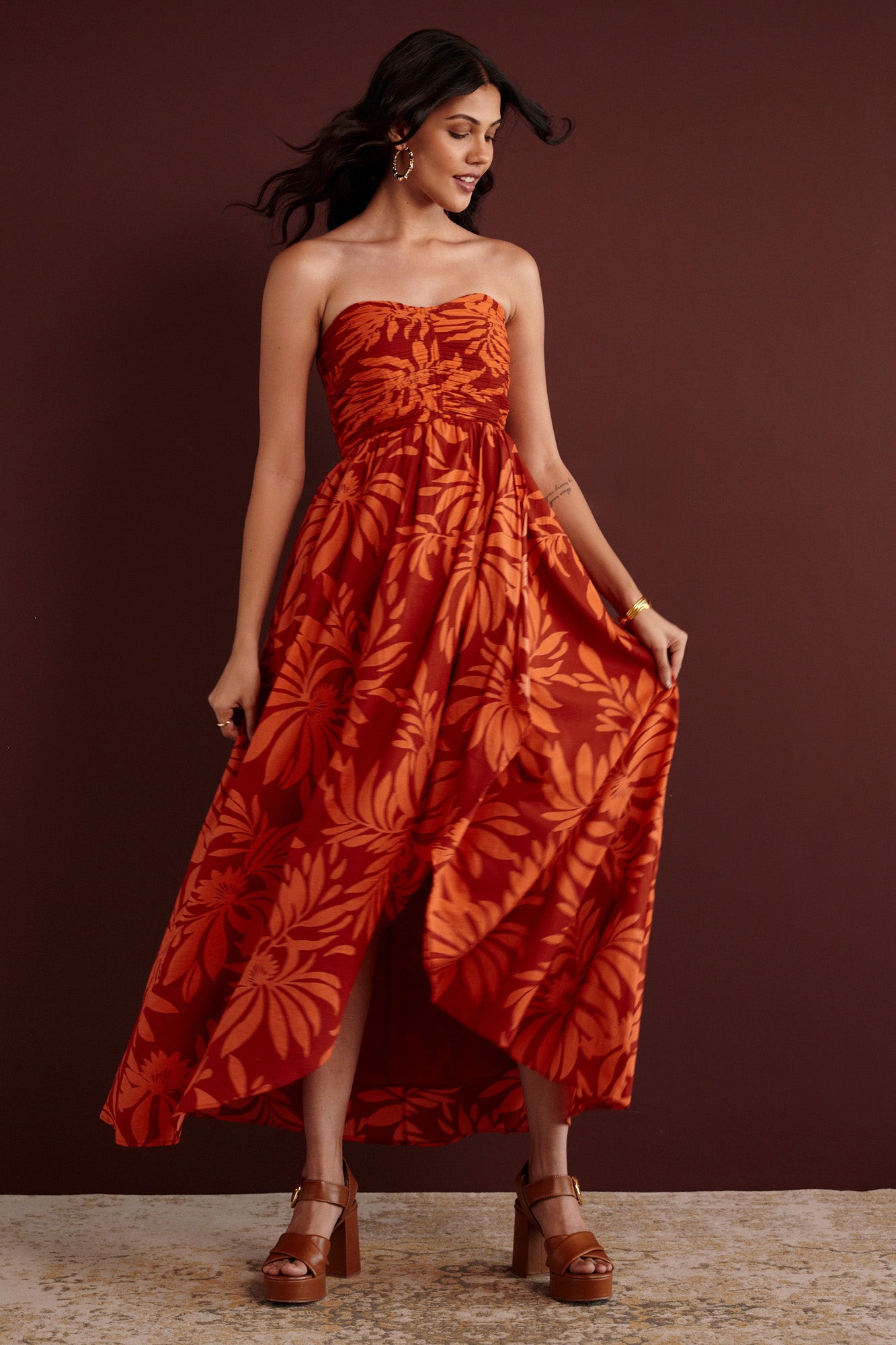 Front view of woman standing. She is wearing the Madison strapless maxi dress in Floral Frond print. It has a ruched bodice, cascading layered skirt, center back zipper, side-seam pockets, and bold Spice color inspired by pomegranate tree embroidery.