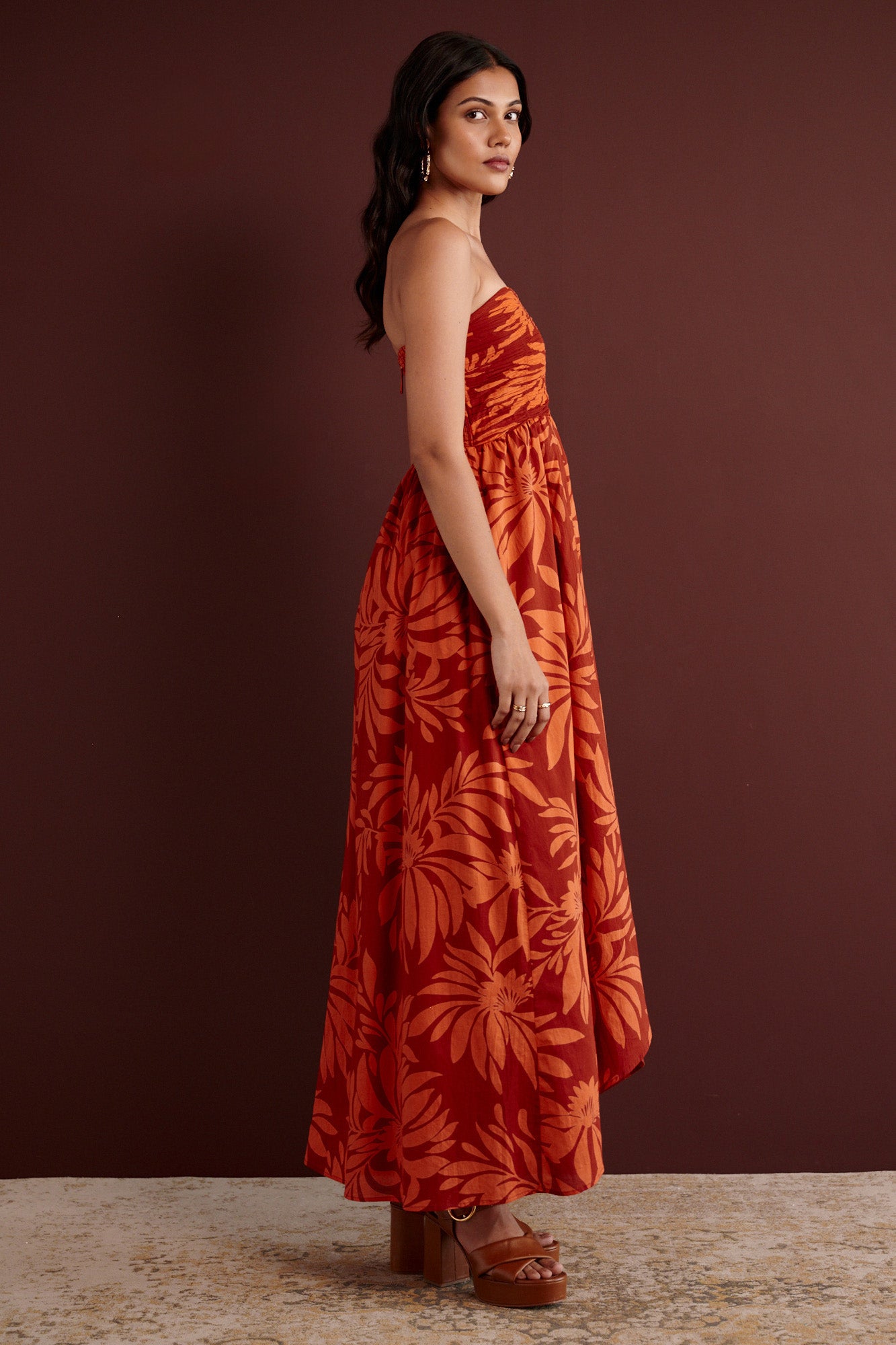 Side view of woman standing. She is wearing the Madison strapless maxi dress in Floral Frond print. It has a ruched bodice, cascading layered skirt, center back zipper, side-seam pockets, and bold Spice color inspired by pomegranate tree embroidery.