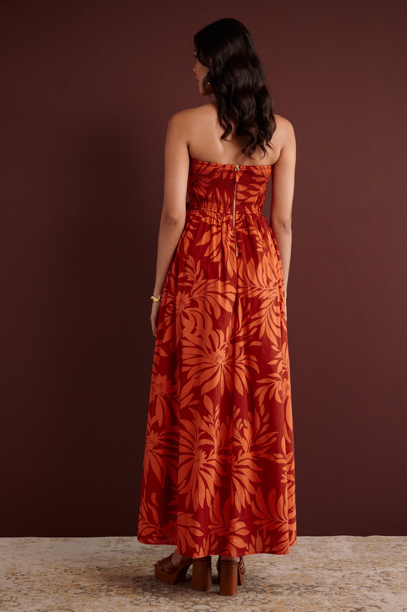 Back view of woman standing. She is wearing the Madison strapless maxi dress in Floral Frond print. It has a ruched bodice, cascading layered skirt, center back zipper, side-seam pockets, and bold Spice color inspired by pomegranate tree embroidery.