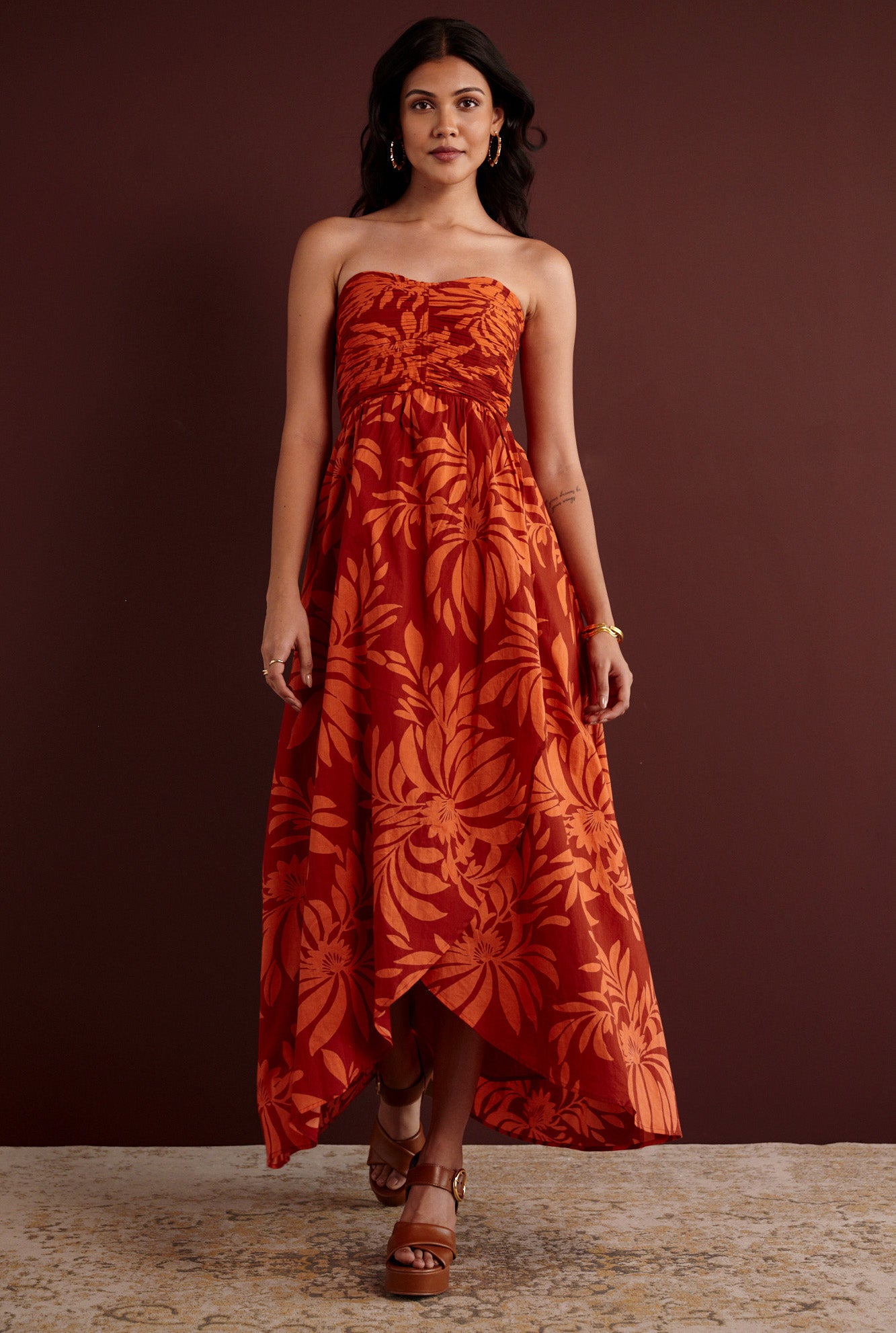 Front view of woman standing. She is wearing the Madison strapless maxi dress in Floral Frond print. It has a ruched bodice, cascading layered skirt, center back zipper, side-seam pockets, and bold Spice color inspired by pomegranate tree embroidery.
