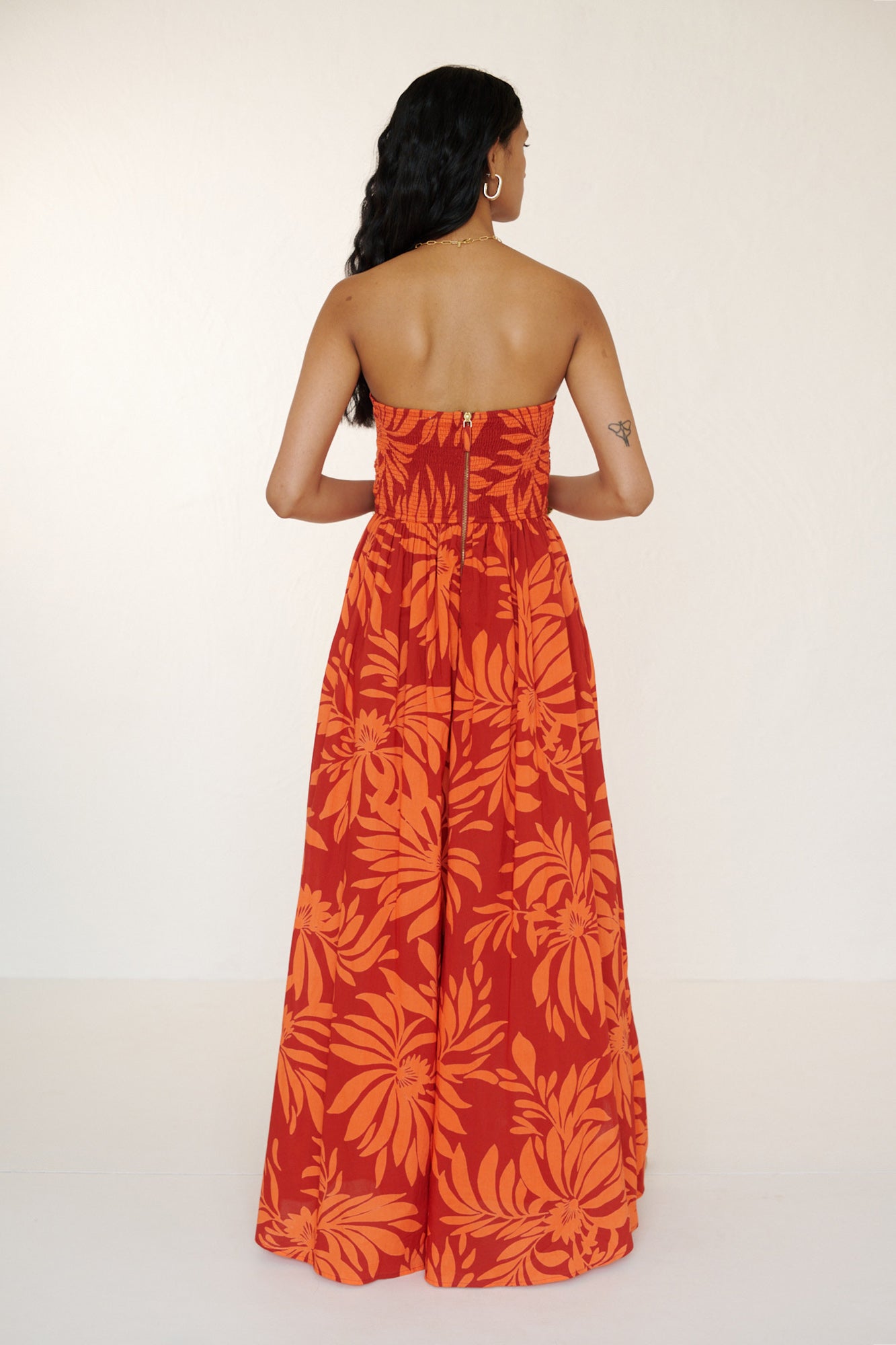 Back view of woman standing. She is wearing the Madison strapless maxi dress in Floral Frond print, with ruched bodice, cascading layered skirt, center back zipper, side-seam pockets, and bold Spice color inspired by pomegranate tree embroidery.