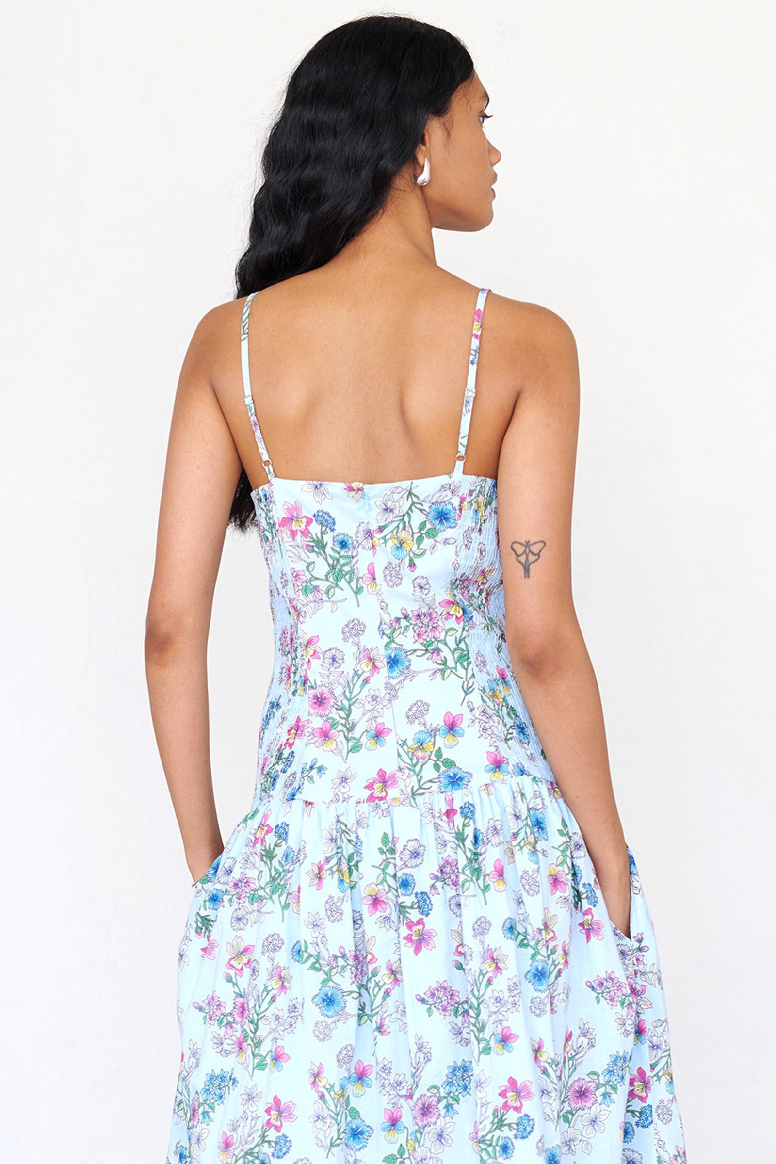 Back view of a woman standing wearing the Maisie sundress in Burdock Imperial print with her hands in pocket. It features smocked sides, adjustable straps, slightly dropped waist and side-seam pockets.