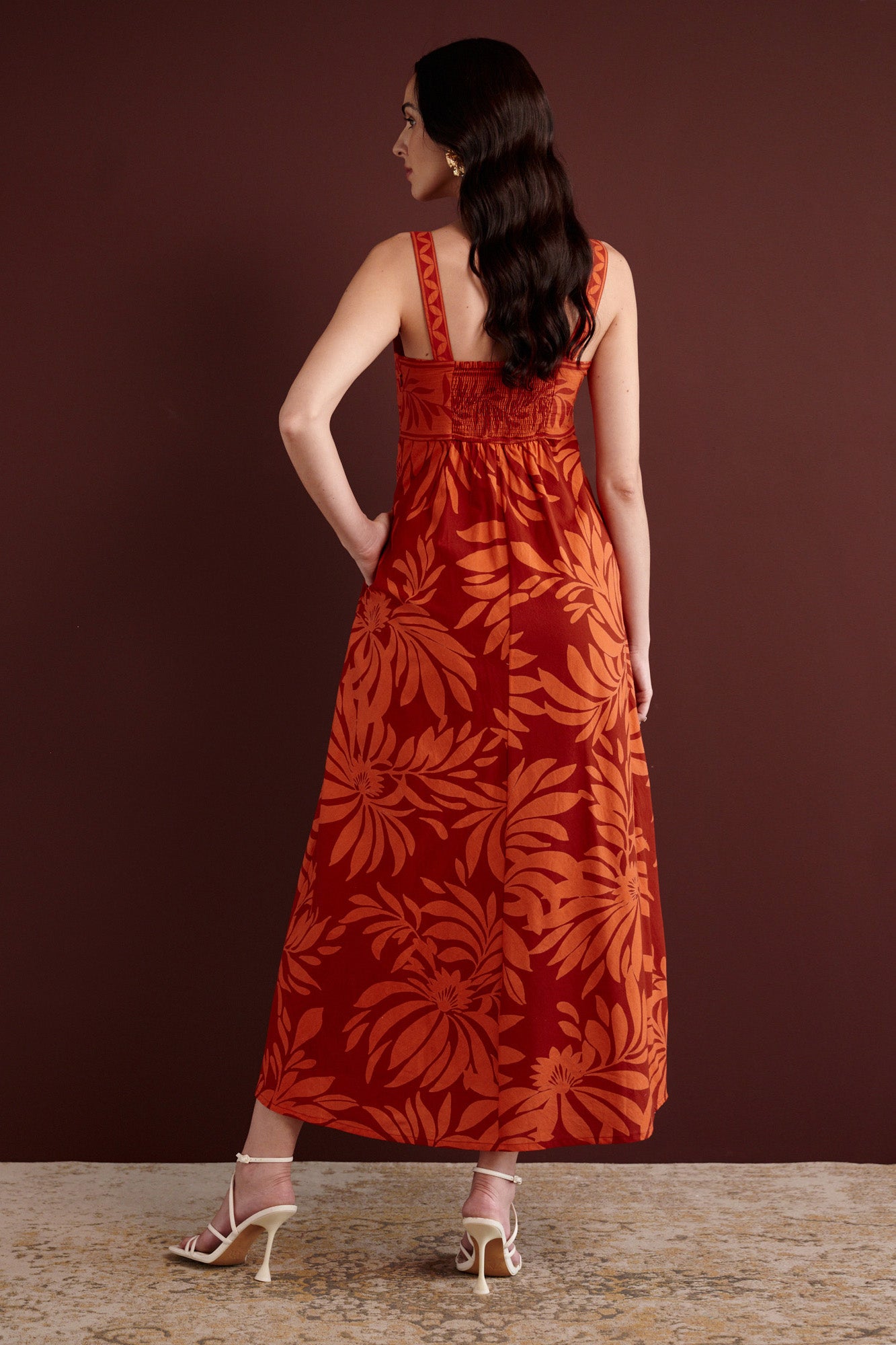 Back view of woman standing. She is wearing the Maria strappy midi dress in Floral Frond print. It is made from lightweight cotton voile, fully lined, with side-seam pockets, in a bold Spice color inspired by pomegranate tree embroidery.