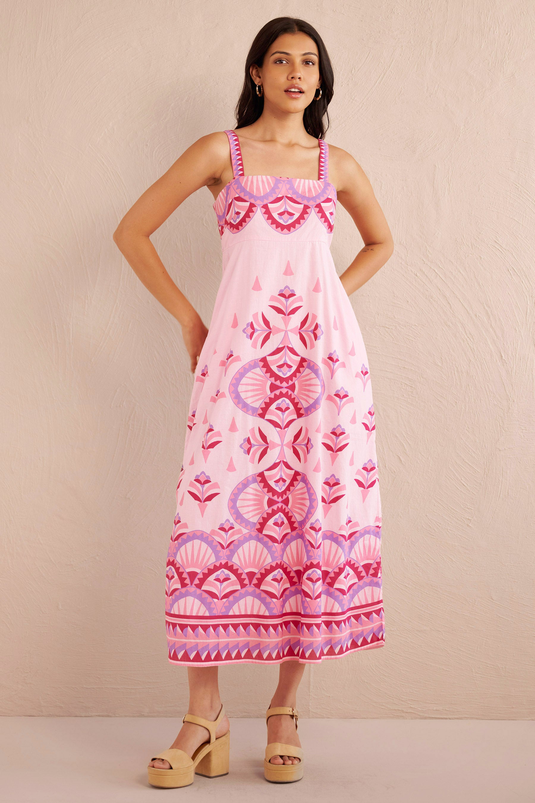Front view of woman wearing a sleeveless pink midi dress with bold geometric amethyst and fuchsia prints, styled with platform sandals.