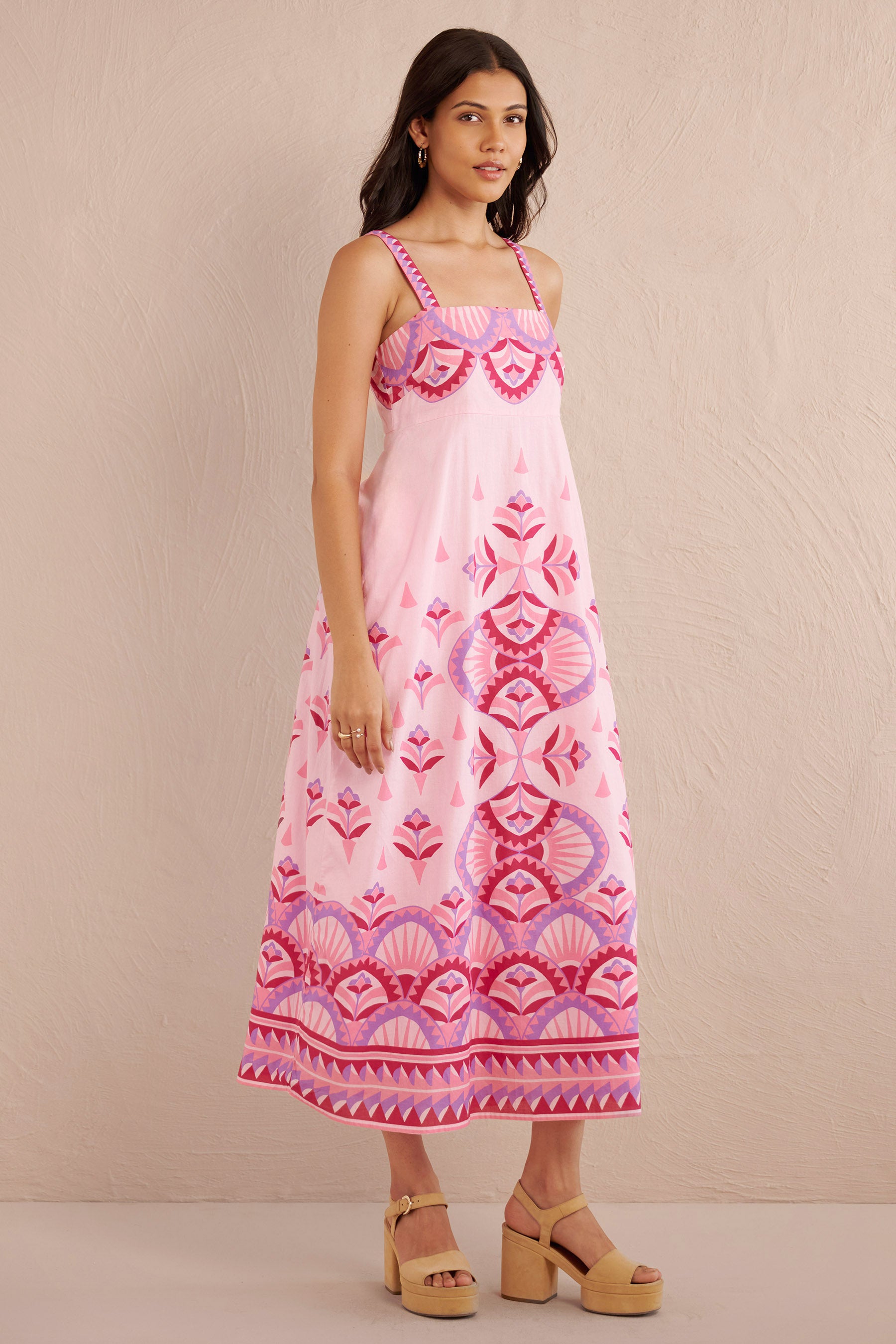 Front side view of woman wearing a sleeveless pink midi dress with bold geometric amethyst and fuchsia prints, styled with platform sandals.