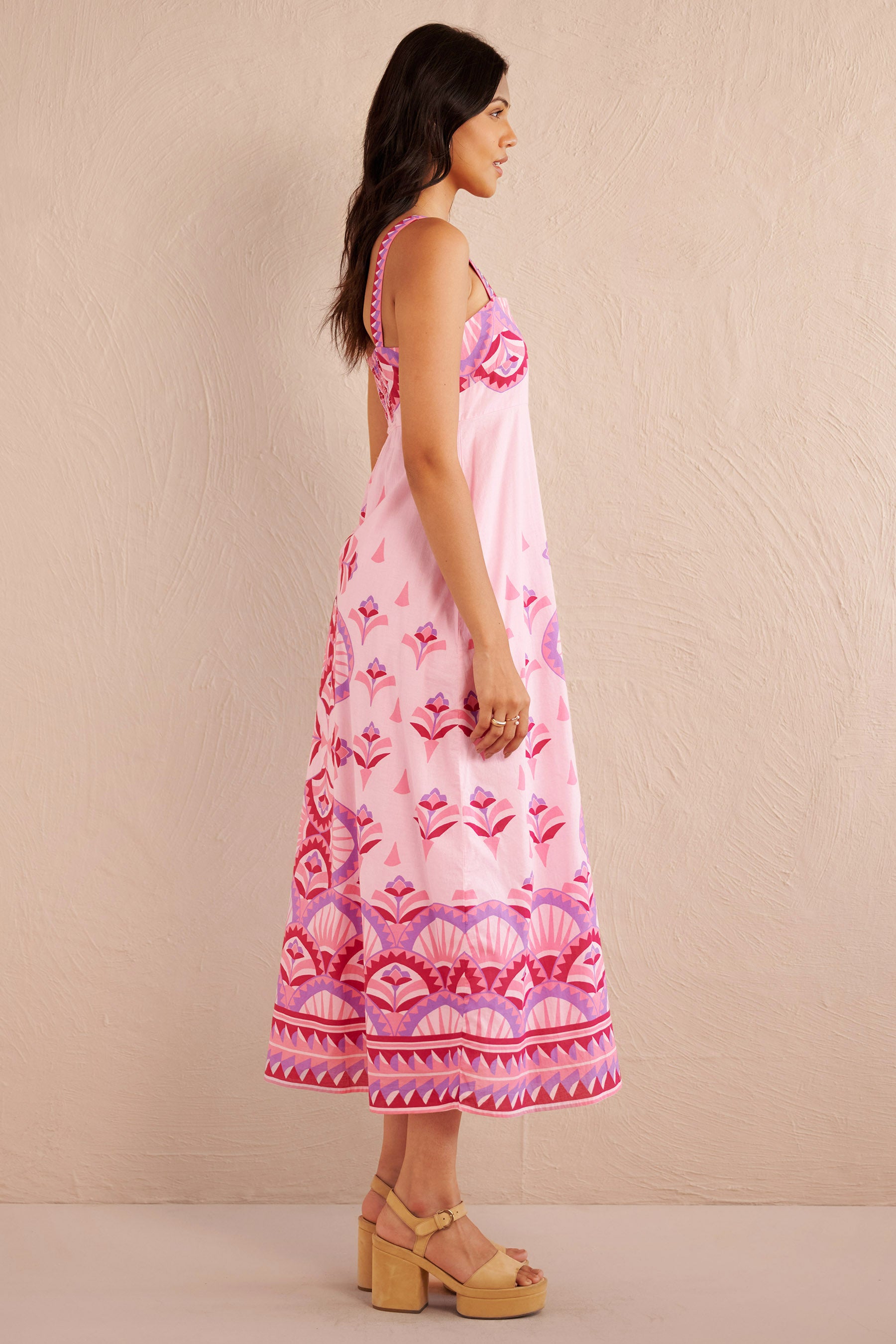 Side view of woman wearing a sleeveless pink midi dress with bold geometric amethyst and fuchsia prints, styled with platform sandals.