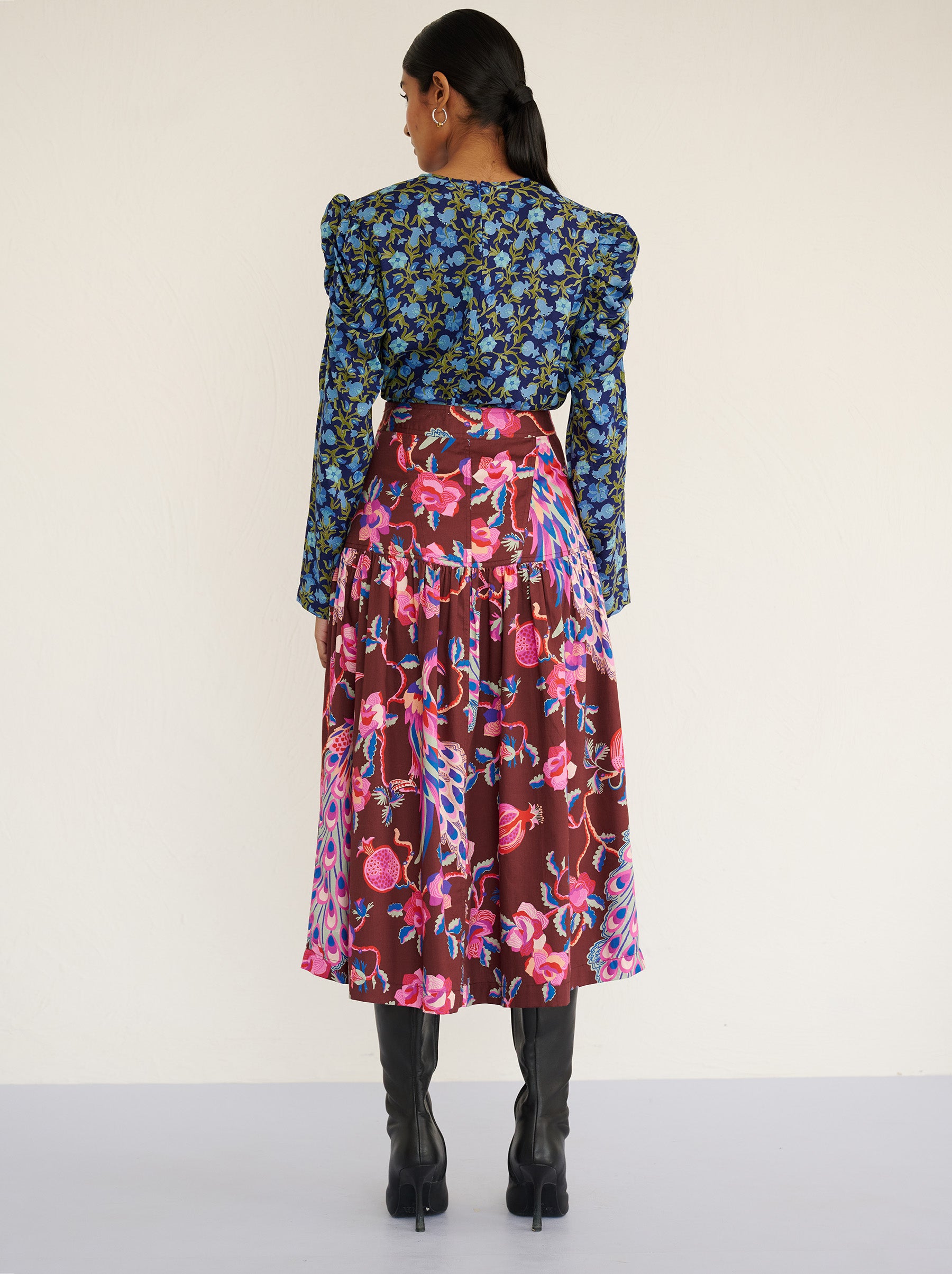 Back view of a model standing. She is wearing the Melodie skirt in cotton sateen with the Bukhara Peacock print in rum color. It is slim at the waist and gathered below for a flattering shape. The skirt is fully lined with side-seam pockets. It is paired with Vincent top in Pomegrante Vine Lapis print with black boots.