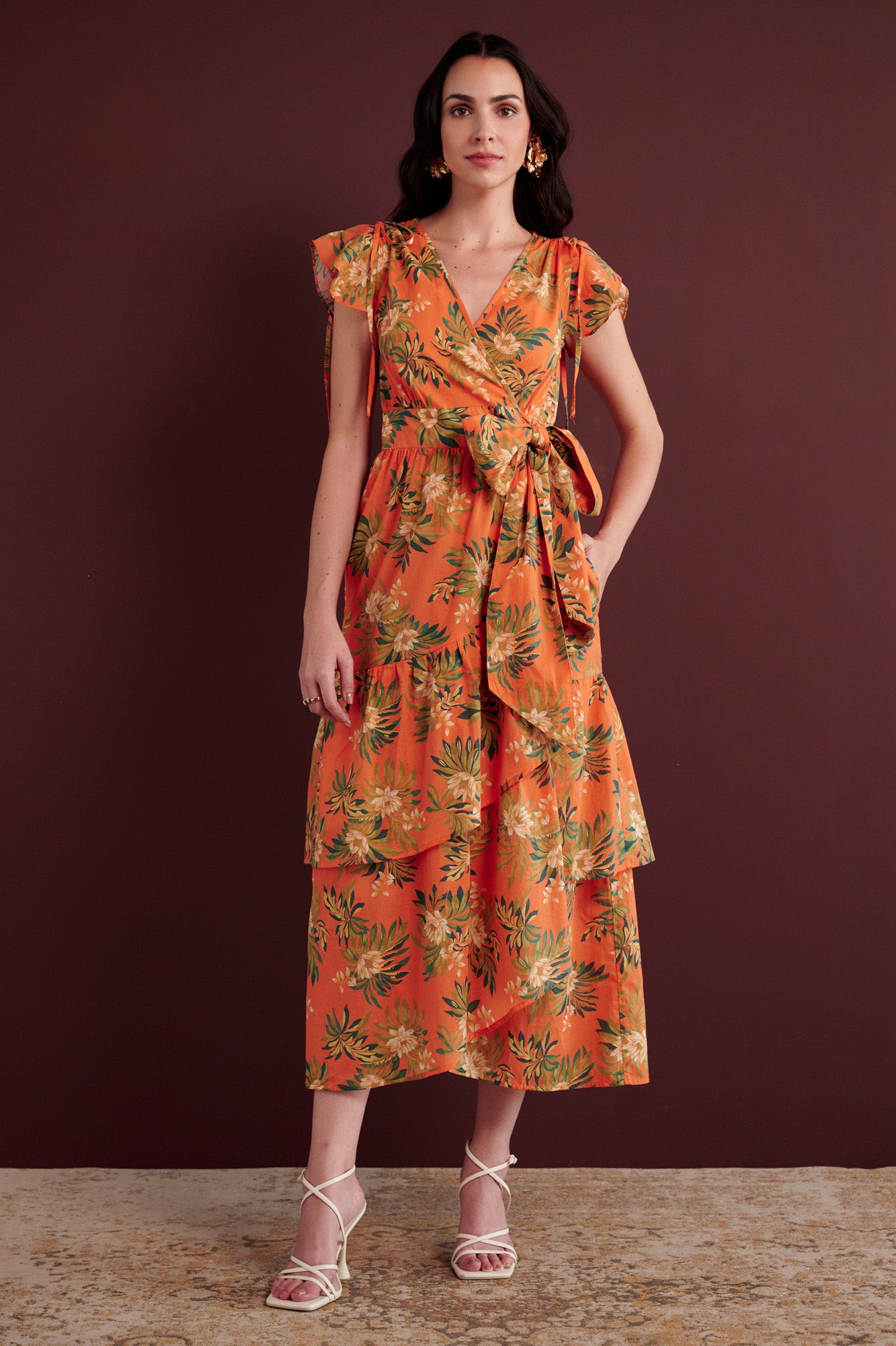 Front view of woman standing with her left hand in pocket. She is wearing the Mercy wrap dress in Artichoke Flower print. It has asymmetrical tiers, adjustable shoulder drawstrings, breezy cotton voile fabric, and side-seam pockets, inspired by 15th-century embroidered tapestry motifs.