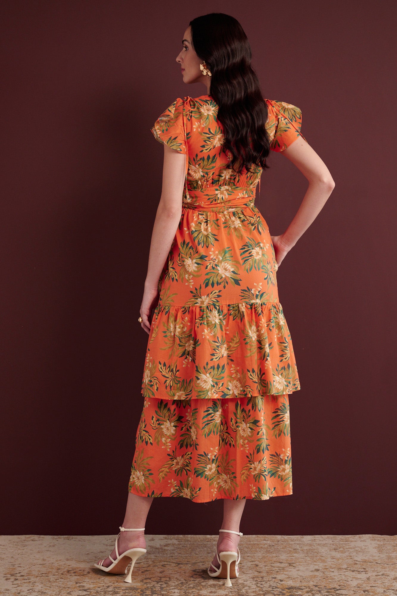 Back view of woman standing with her left hand in pocket. She is wearing the Mercy wrap dress in Artichoke Flower print. It has asymmetrical tiers, adjustable shoulder drawstrings, breezy cotton voile fabric, and side-seam pockets, inspired by 15th-century embroidered tapestry motifs.
