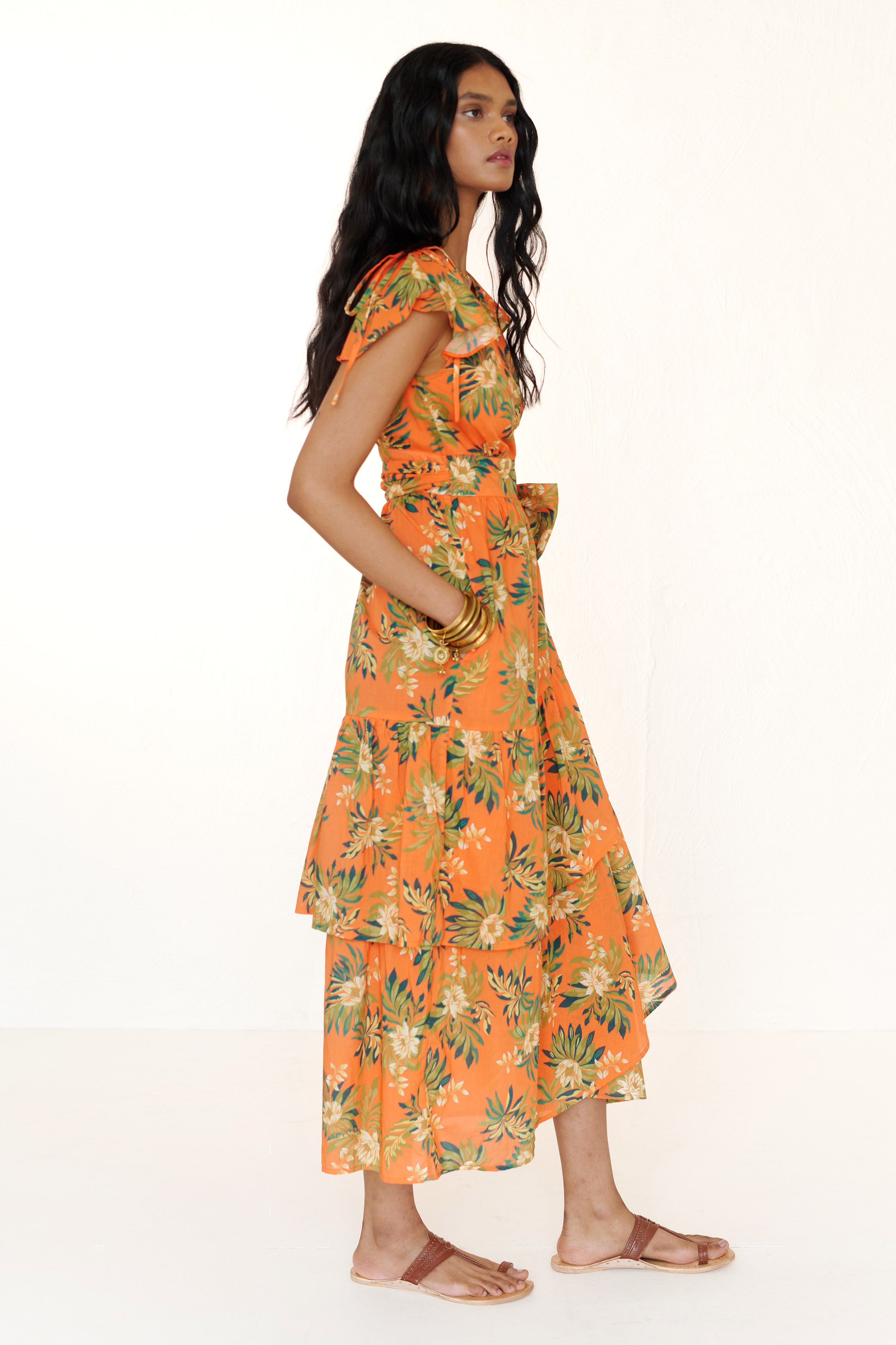 Side view of woman standing. She is wearing the Mercy wrap dress in Artichoke Flower print. It has asymmetrical tiers, adjustable shoulder drawstrings, breezy cotton voile fabric, and side-seam pockets, inspired by 15th-century embroidered tapestry motifs.
