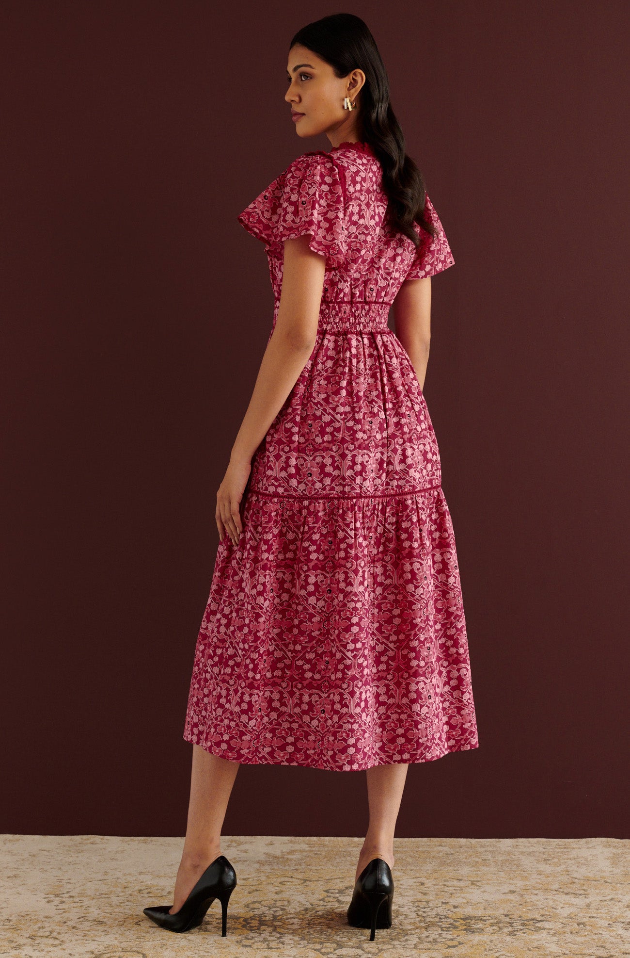 Back view of woman standing. She is wearing the Naxos midi dress in Ottoman print, with plunging V-neck trimmed in red lace, lace insets at waist and skirt, flutter sleeves, smooth cotton cambric, fully lined construction, and side-seam pockets.