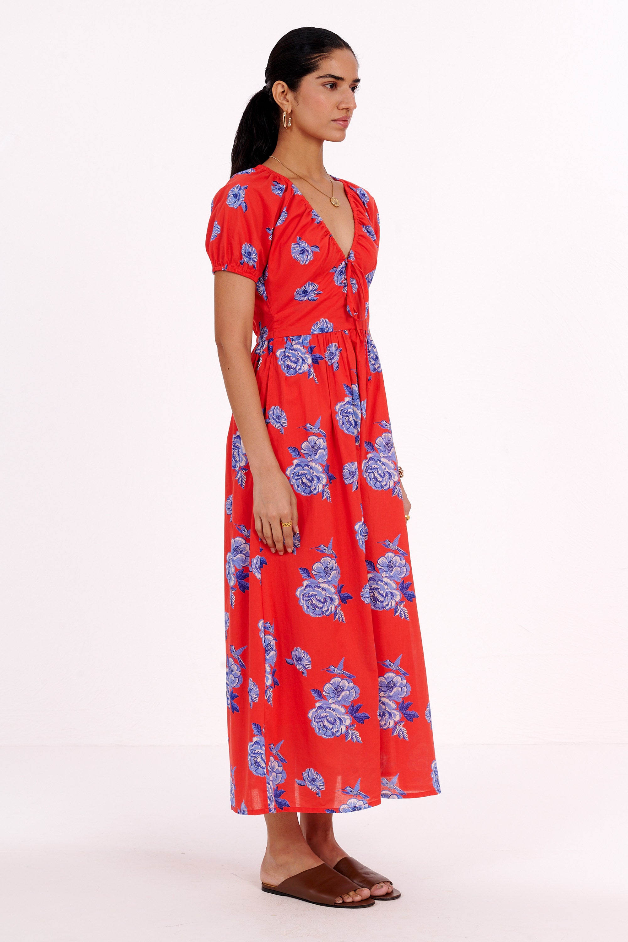 Side view of woman standing. She is wearing the Norma dress in the Eliza Rose print, featuring a drawstring neckline, princess-seam waist, elastic sleeves, and floral motifs with blue roses and hummingbirds.