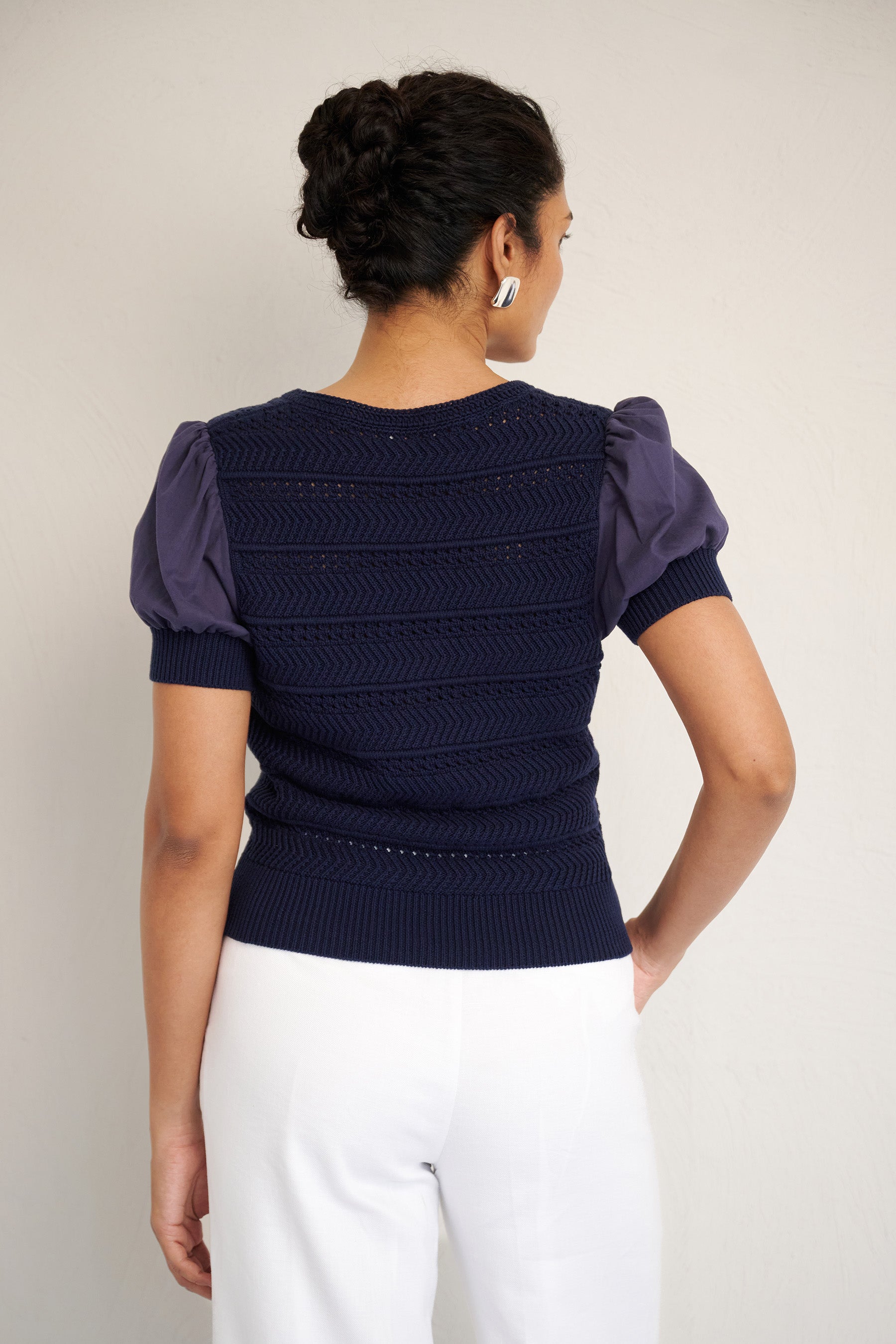 Back view of woman standing. She is wearing Patti crew knit in Oxford blue color. It is a 100% cotton with puffed cotton voile sleeves, crochet trim, and textured knit body.