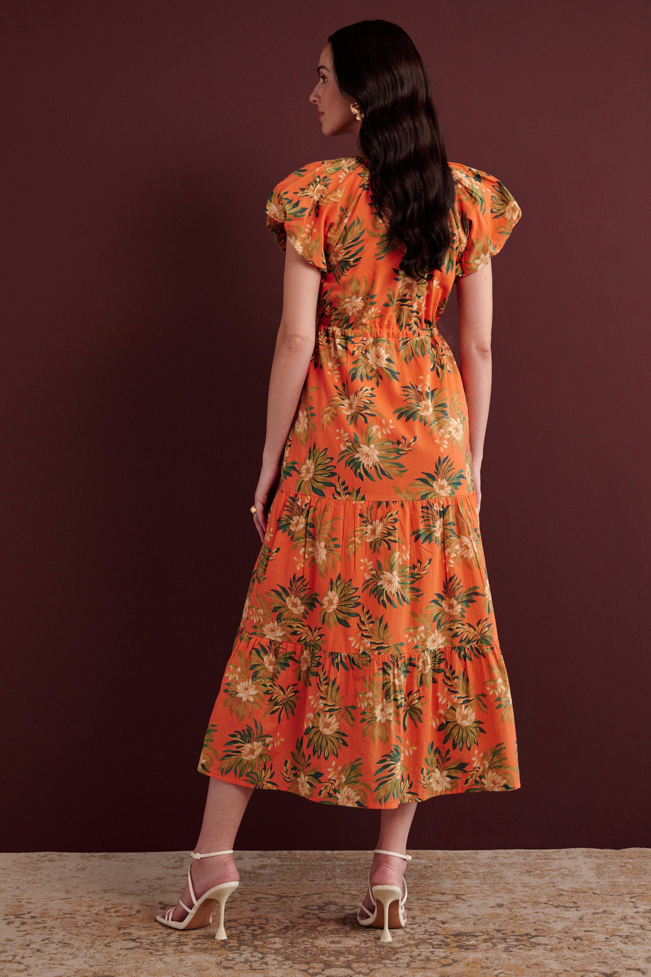 Back view of woman standing. She is wearing the Poppy dress in Artichoke Flower print. It has a raglan blouson sleeves, front button placket, drawstring waist, flowing skirt, cap sleeves, fully lined construction, and side-seam pockets.