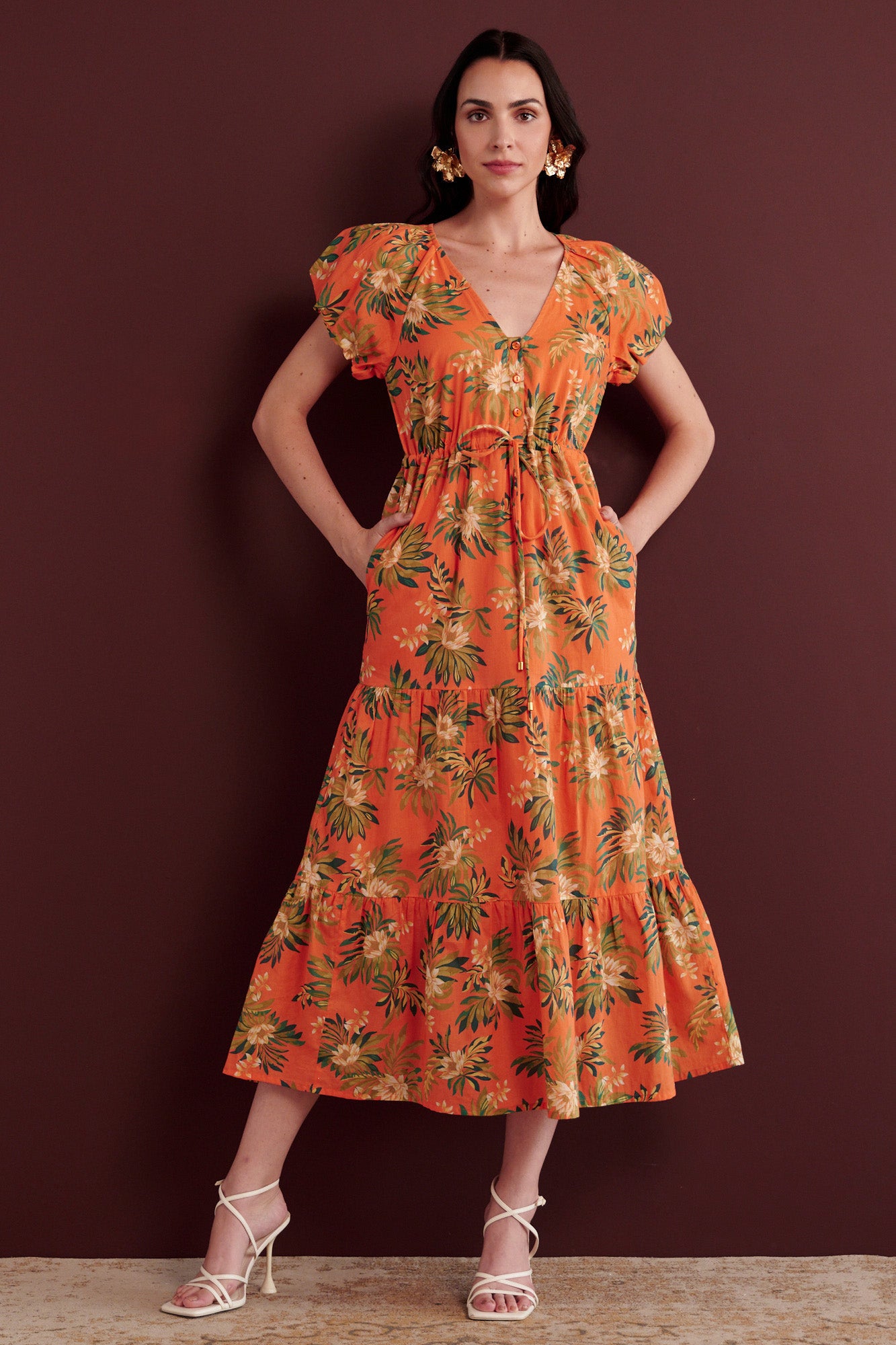 Front view of woman standing with her hands in pocket. She is wearing the Poppy dress in Artichoke Flower print. It has a raglan blouson sleeves, front button placket, drawstring waist, flowing skirt, cap sleeves, fully lined construction, and side-seam pockets.