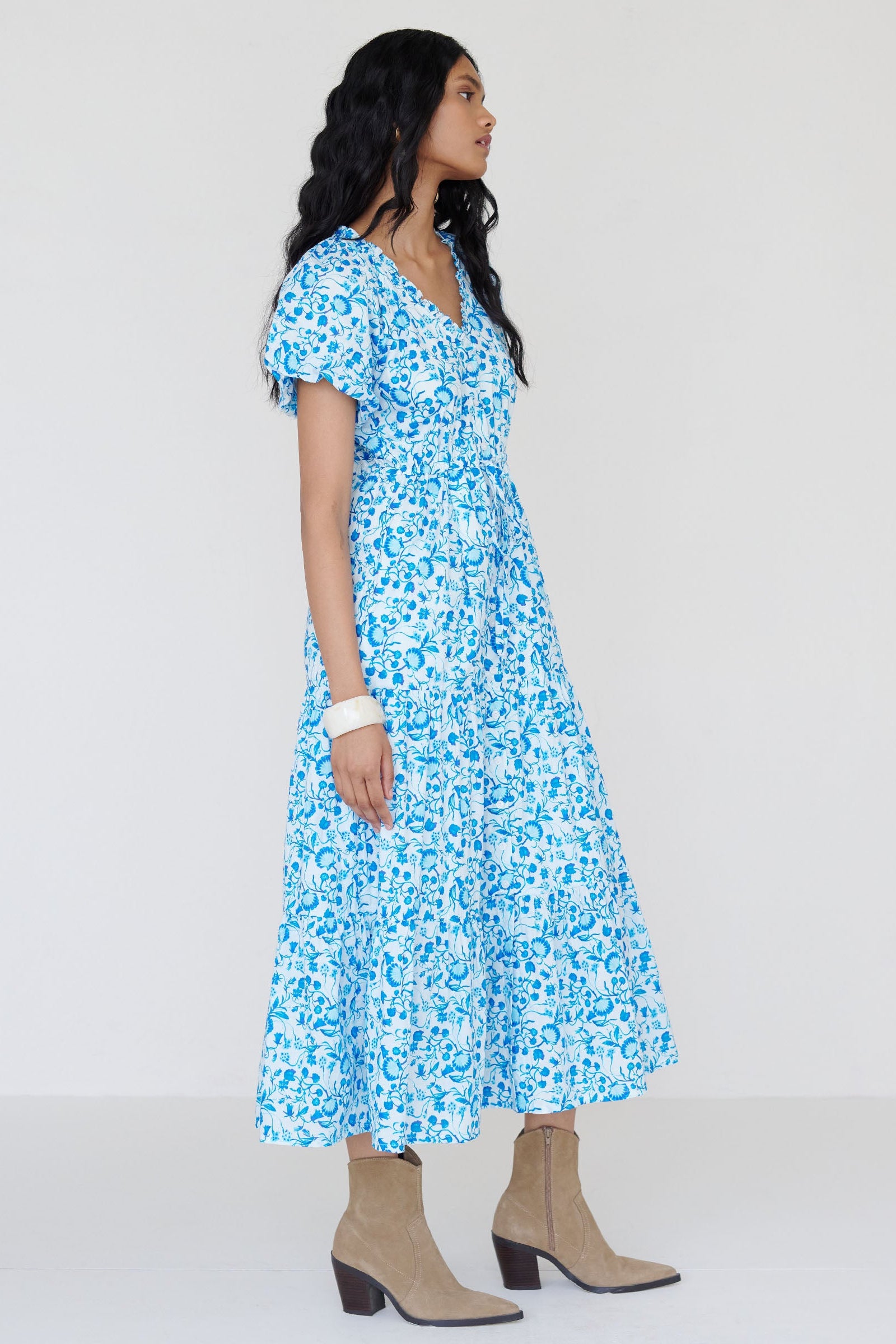 Side view of a woman standing. She is wearing the Poppy dress in Clam Garland print. It features raglan blouson sleeves, a button-front placket, a long flowing skirt, and a drawstring waist.