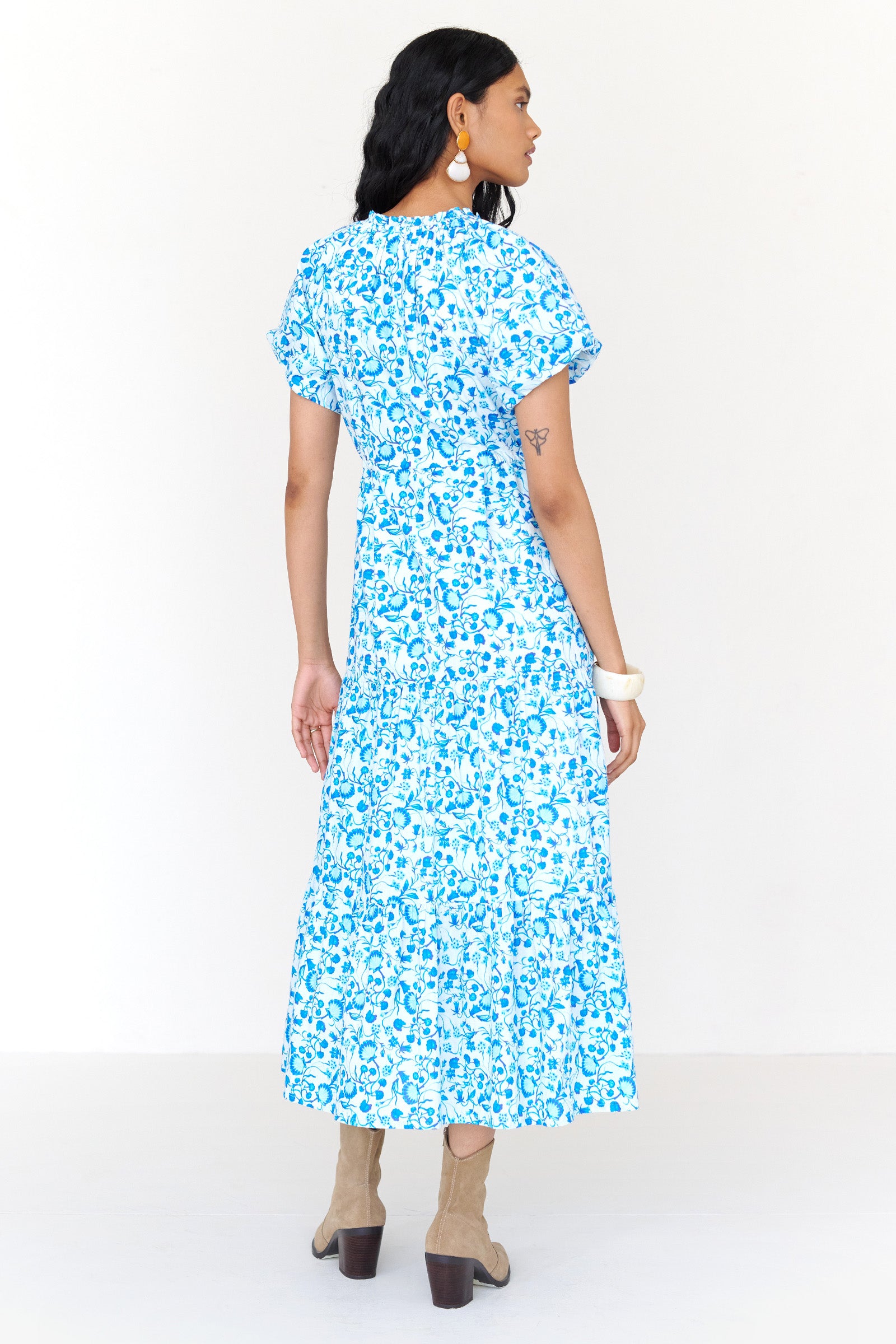 Back view of a woman standing wearing the Poppy dress in Clam Garland print. It features raglan blouson sleeves, front button placket, drawstring waist, flowing skirt and side-seam pockets.