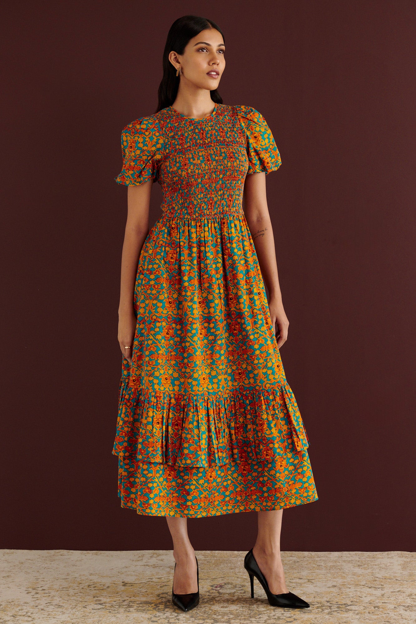 Front side view of woman standing. She is wearing the Quant midi dress in Ottoman print. It has a smocked fitted bodice, subtly puffed sleeves, full tiered skirt, fully lined construction, and side-seam pockets.