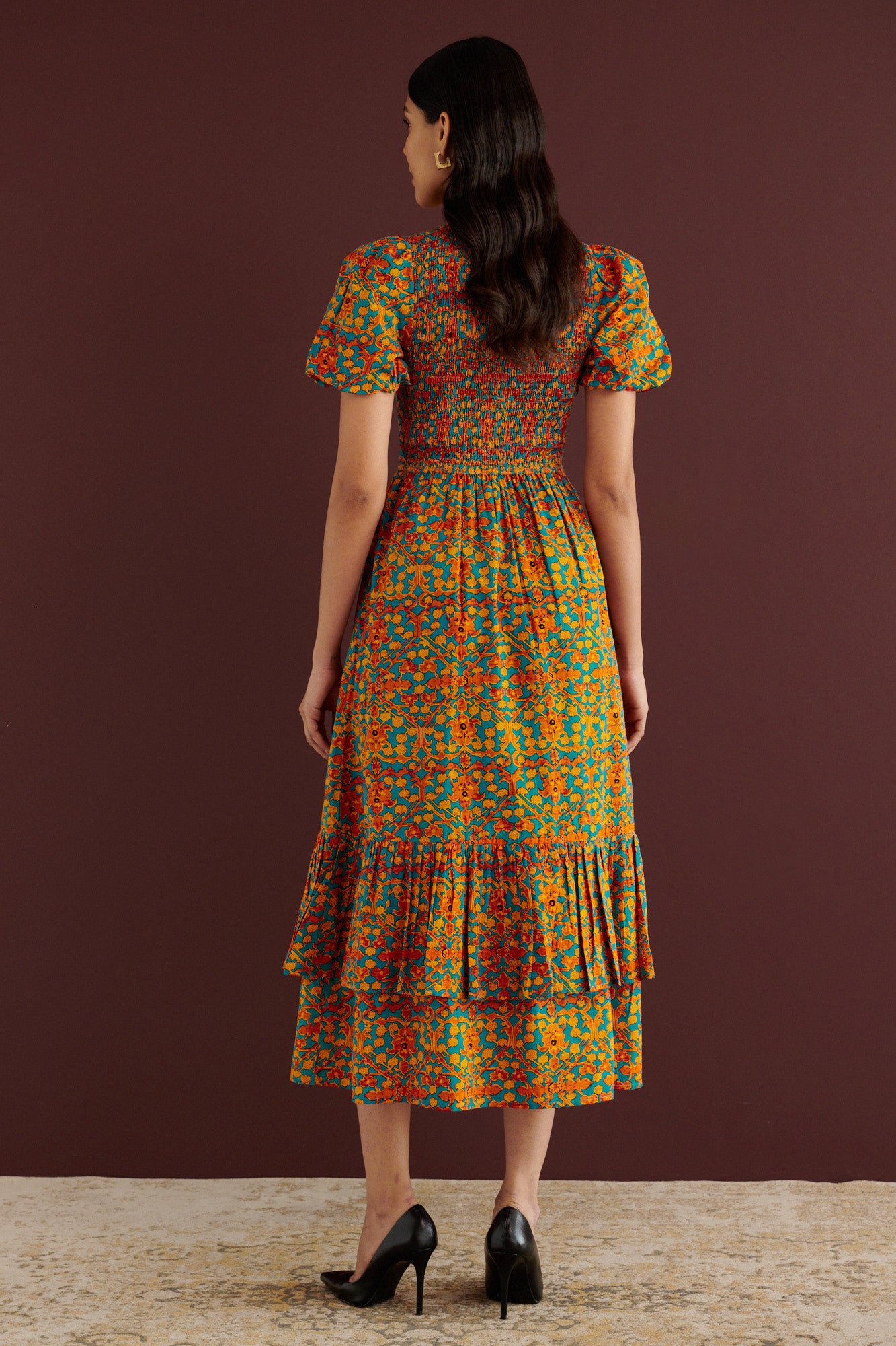Back view of woman standing. She is wearing the Quant midi dress in Ottoman print. It has a smocked fitted bodice, subtly puffed sleeves, full tiered skirt, fully lined construction, and side-seam pockets.