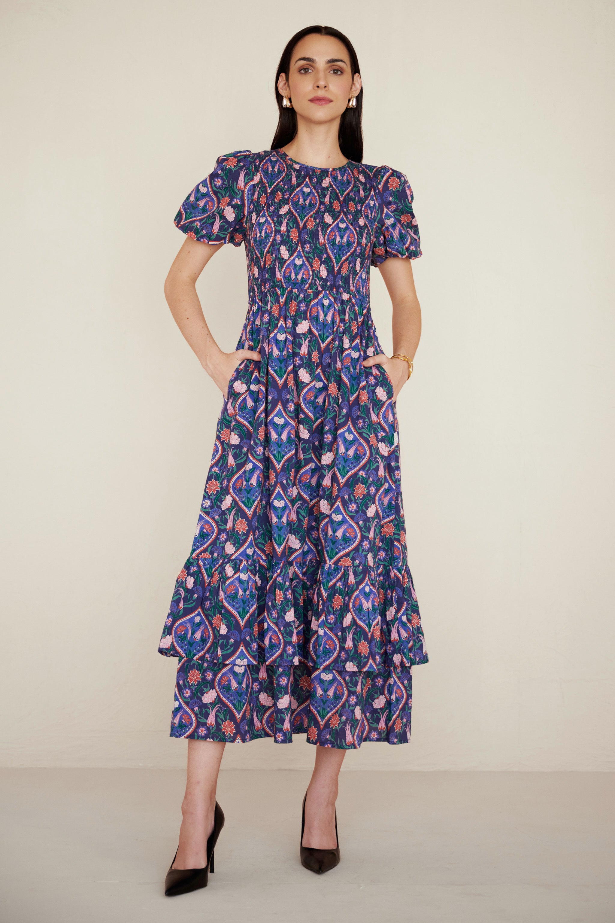 Front view of woman standing. She is wearing a Quant dress in Persian Tile print in Lapis color with her both hands in pocket. It is a midi dress with smocked bodice, puff sleeves, and tiered skirt, featuring a floral tile print inspired by Uzbek design.