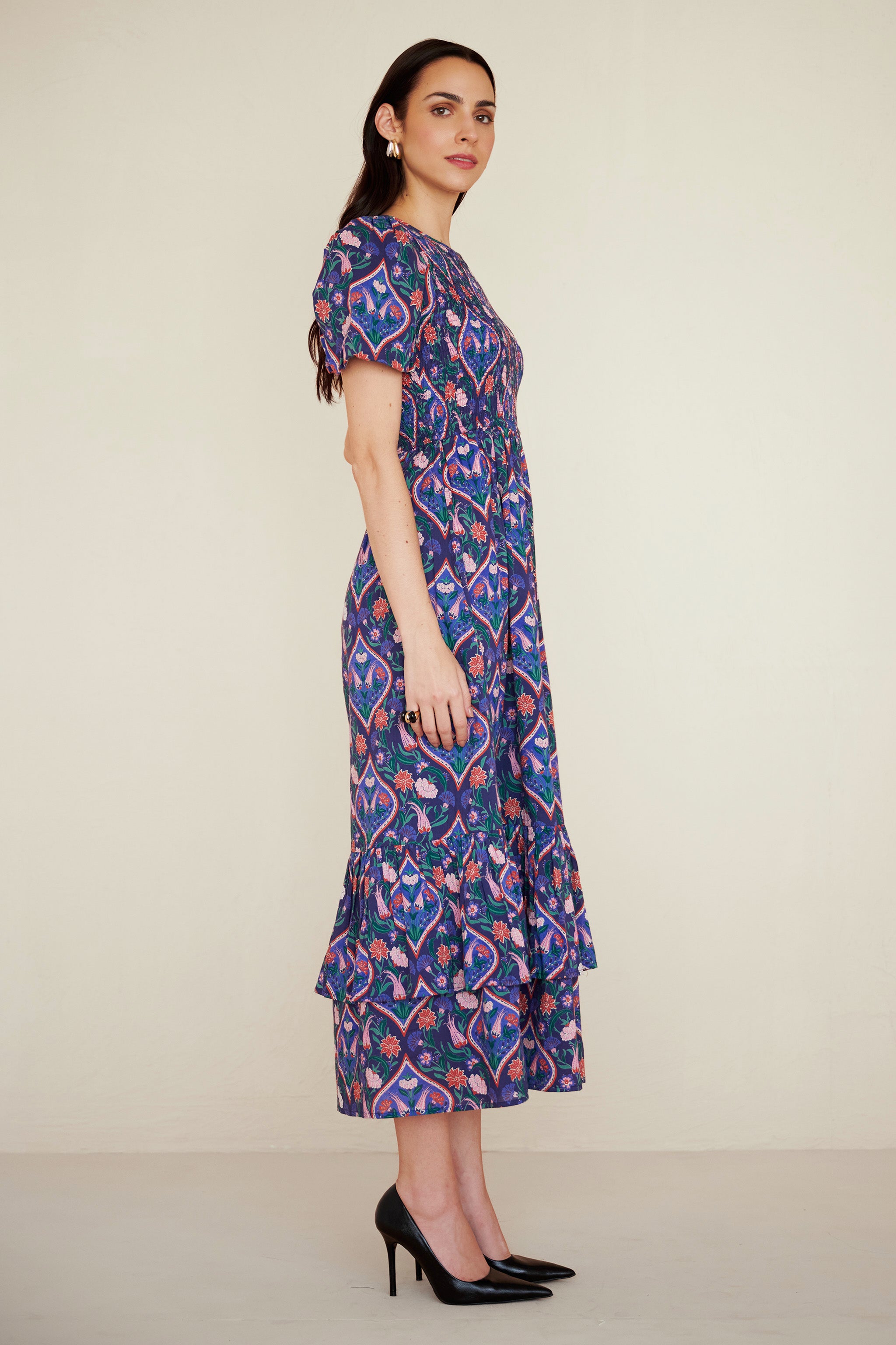 Side view of woman wearing a midi dress with smocked bodice, puff sleeves, and tiered skirt, featuring a floral tile print inspired by Uzbek design.
