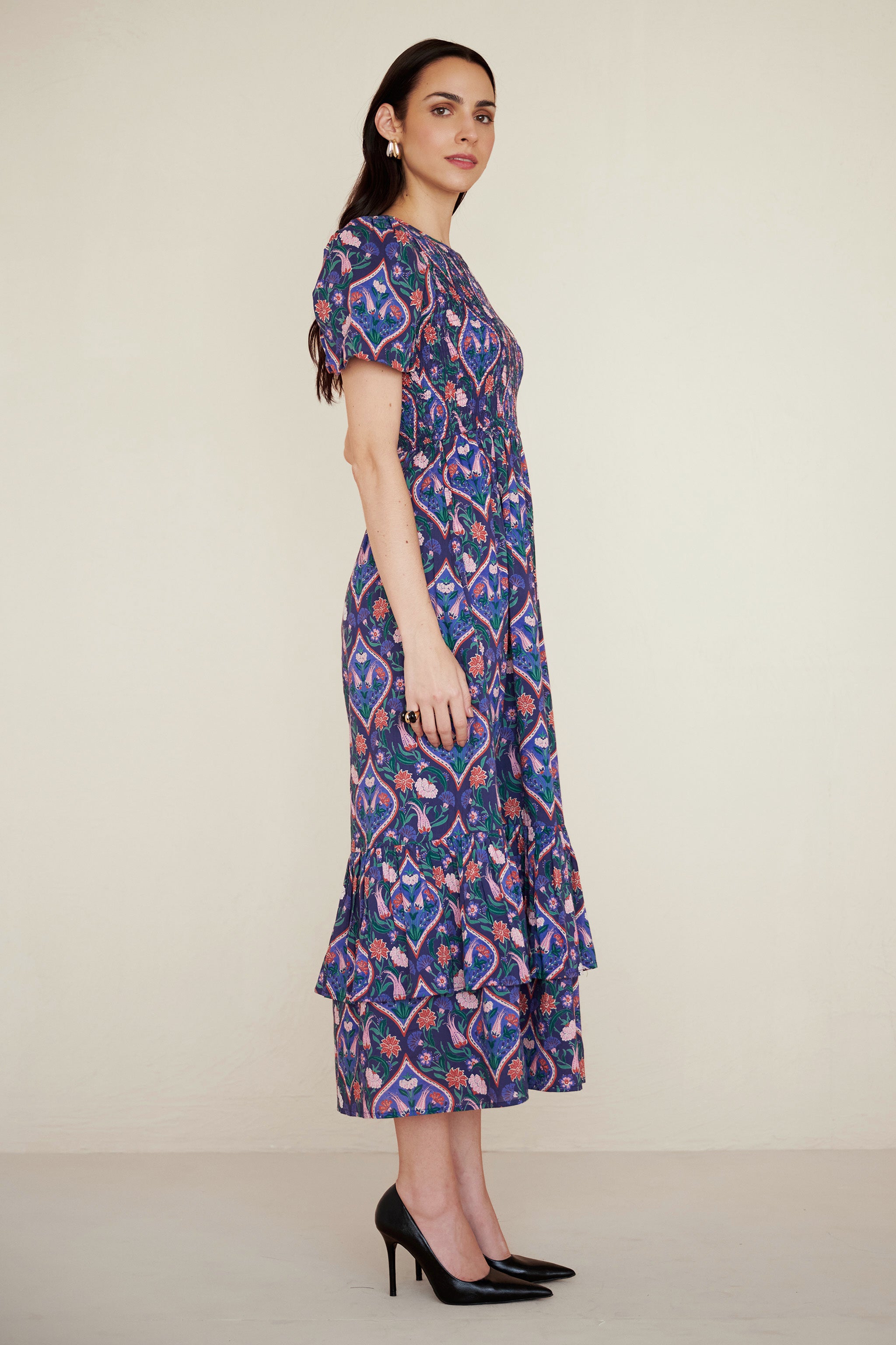 Side view of woman standing. She is woman wearing a Quant dress in Persian Tile print in Lapis color. It is a midi dress with smocked bodice, puff sleeves, and tiered skirt, featuring a floral tile print inspired by Uzbek design.