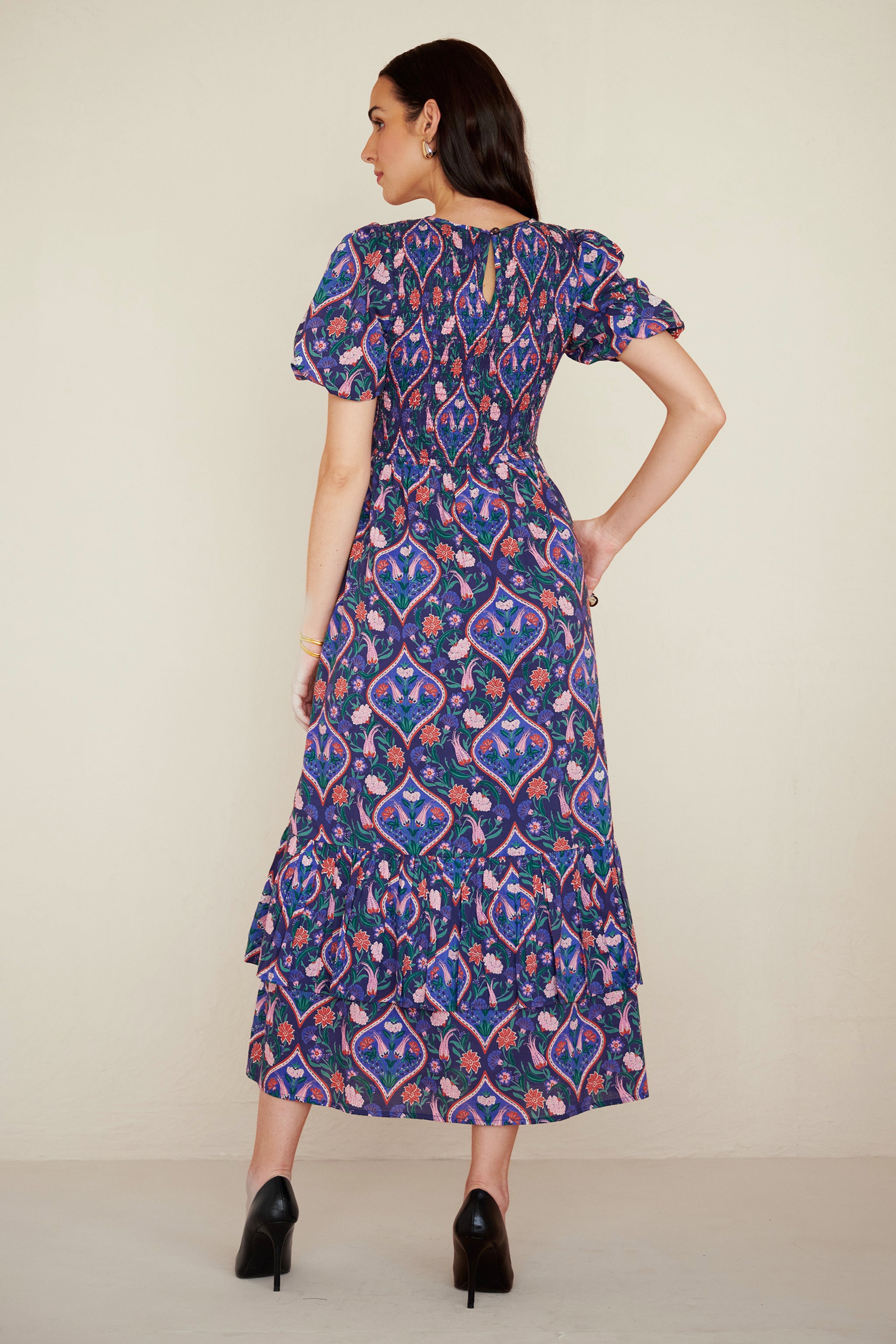 Back view of woman wearing a midi dress with smocked bodice, puff sleeves, and tiered skirt, featuring a floral tile print inspired by Uzbek design.
