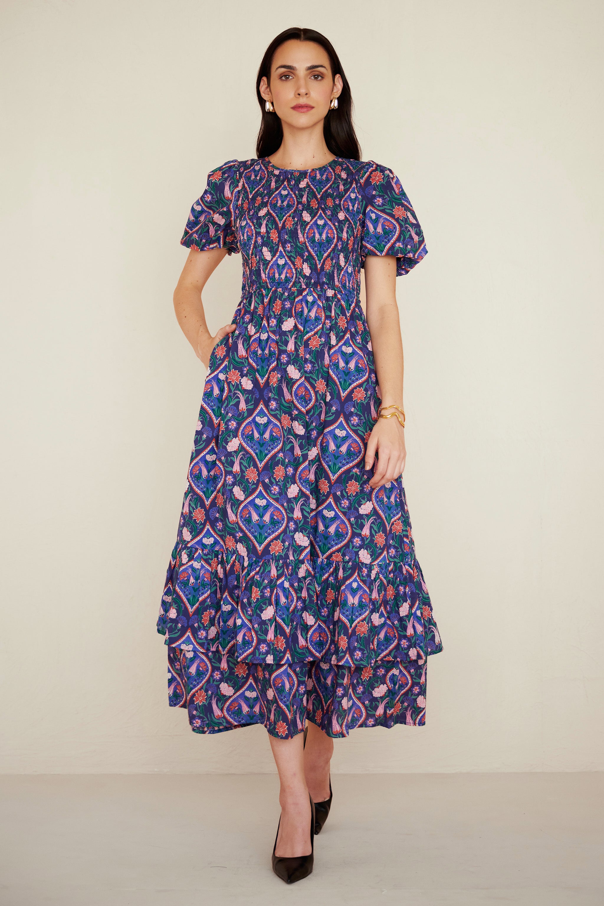 Front view of woman wearing a midi dress with smocked bodice, puff sleeves, and tiered skirt, featuring a floral tile print inspired by Uzbek design.
