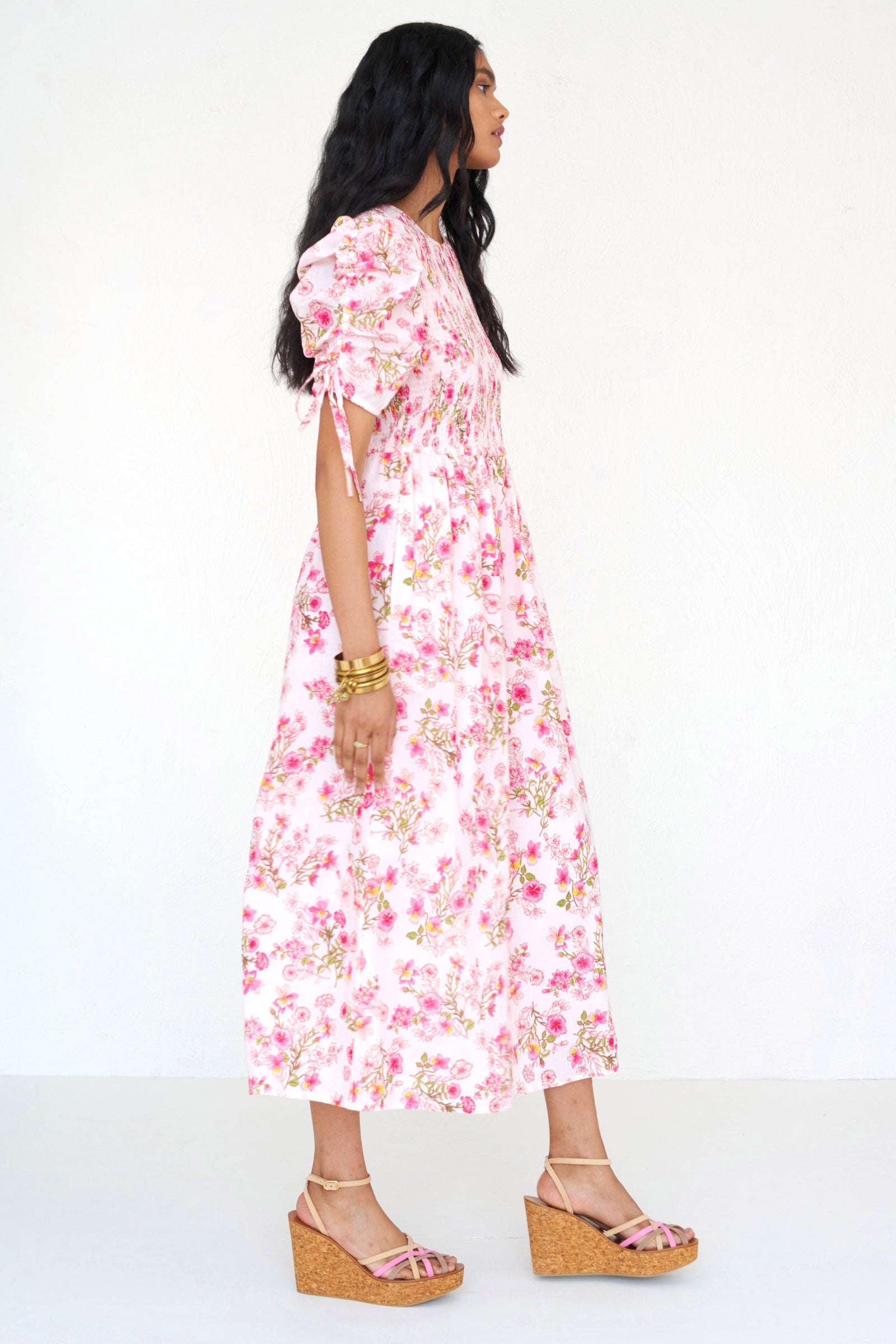 Side view of a woman standing wearing the Queenie midi dress in Burdock floral print, featuring a smocked bodice, drawstring sleeves, full skirt & pockets.
