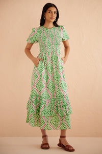 Front view of woman wearing the Quant midi dress in Athena Heart print, featuring a smocked bodice, puffed sleeves, tiered full skirt, lace trim, and side pockets in a green and pink floral-heart motif. Front view of woman wearing the Quant midi dress in Athena Heart print, featuring a smocked bodice, puffed sleeves, tiered full skirt, lace trim, and side pockets in a green and pink floral-heart motif.