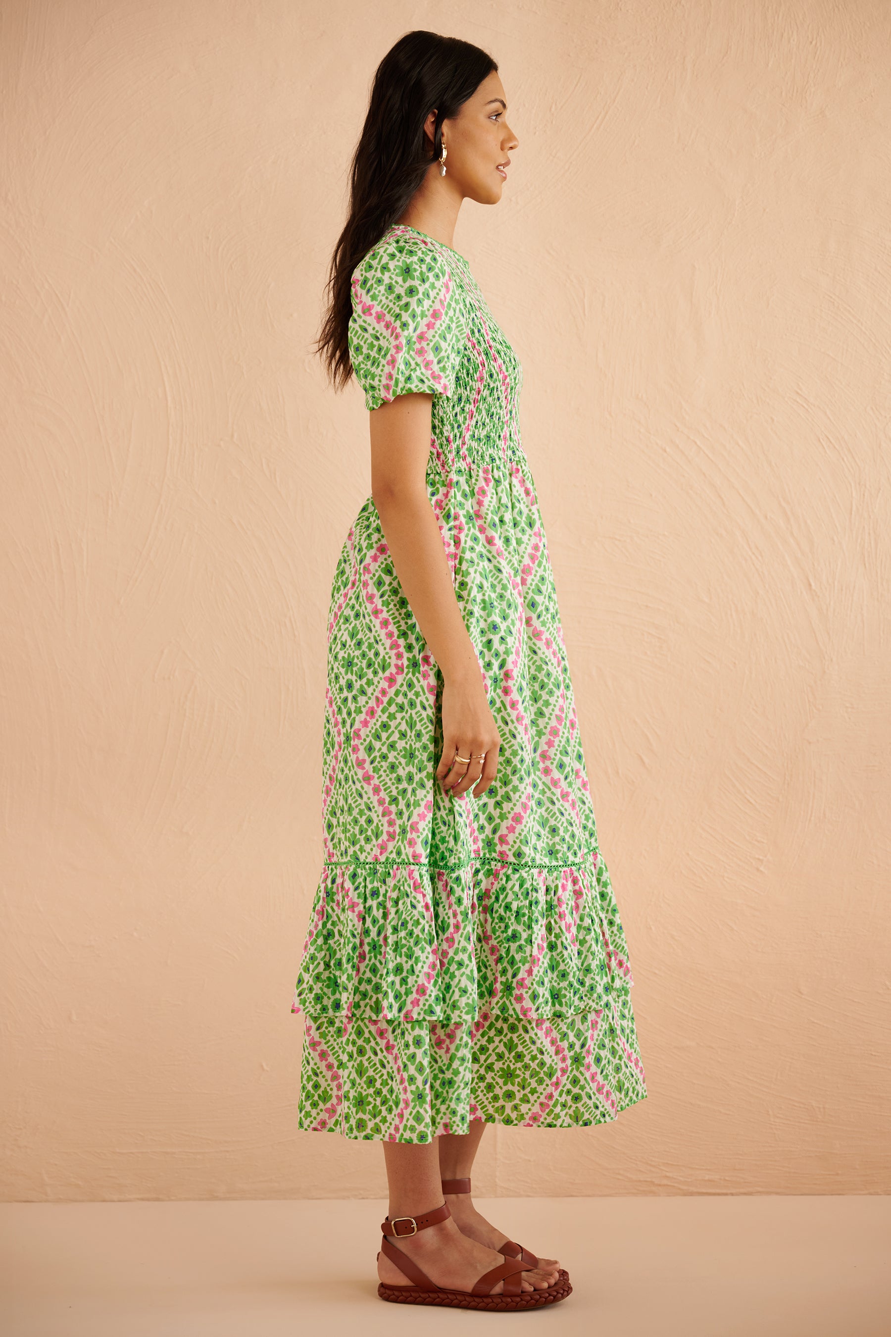 Side view of woman wearing the Quant midi dress in Athena Heart print, featuring a smocked bodice, puffed sleeves, tiered full skirt, lace trim, and side pockets in a green and pink floral-heart motif. Side view of woman wearing the Quant midi dress in Athena Heart print, featuring a smocked bodice, puffed sleeves, tiered full skirt, lace trim, and side pockets in a green and pink floral-heart motif.