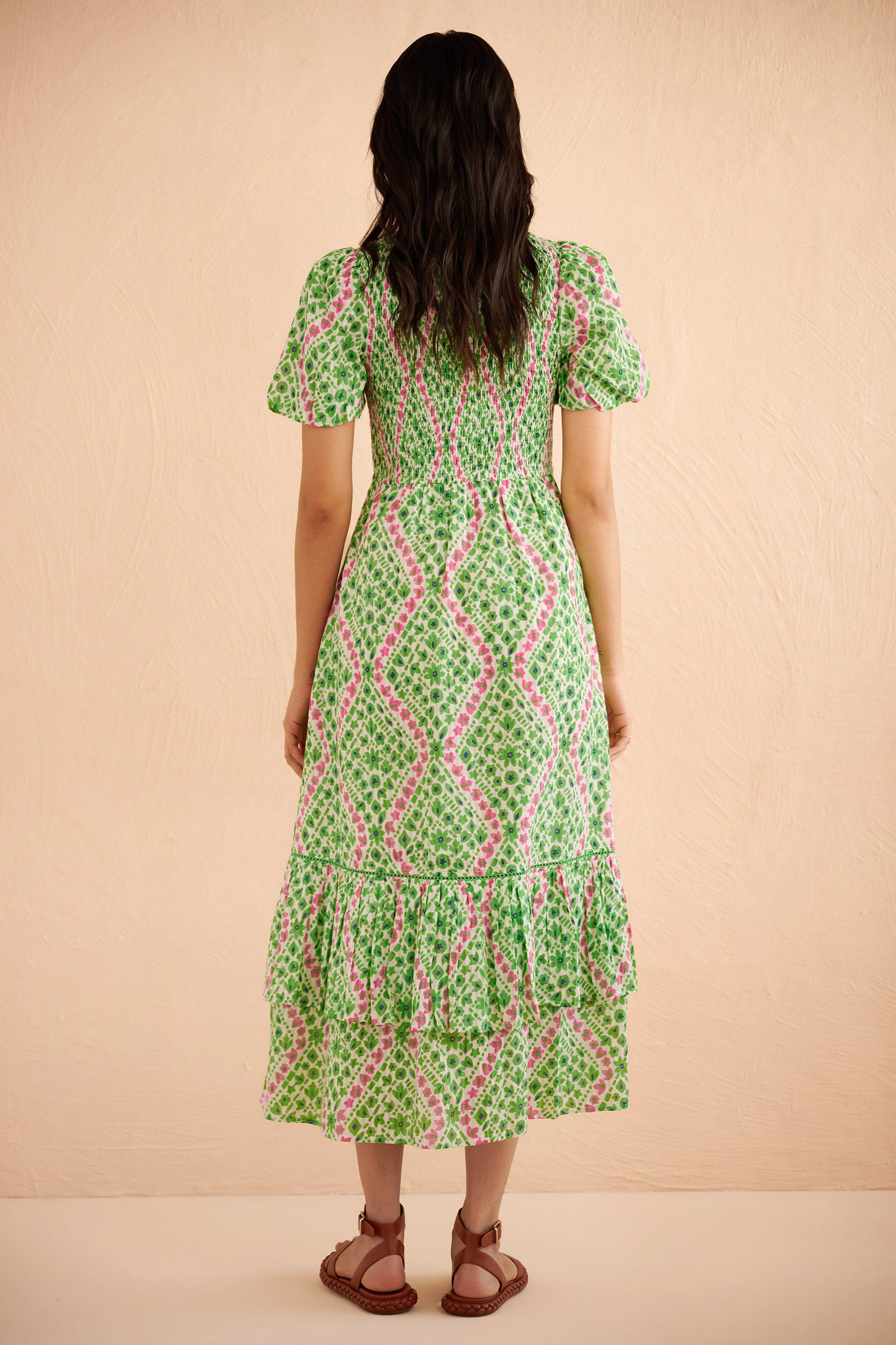 Back view of woman wearing the Quant midi dress in Athena Heart print, featuring a smocked bodice, puffed sleeves, tiered full skirt, lace trim, and side pockets in a green and pink floral-heart motif. Back view of woman wearing the Quant midi dress in Athena Heart print, featuring a smocked bodice, puffed sleeves, tiered full skirt, lace trim, and side pockets in a green and pink floral-heart motif.