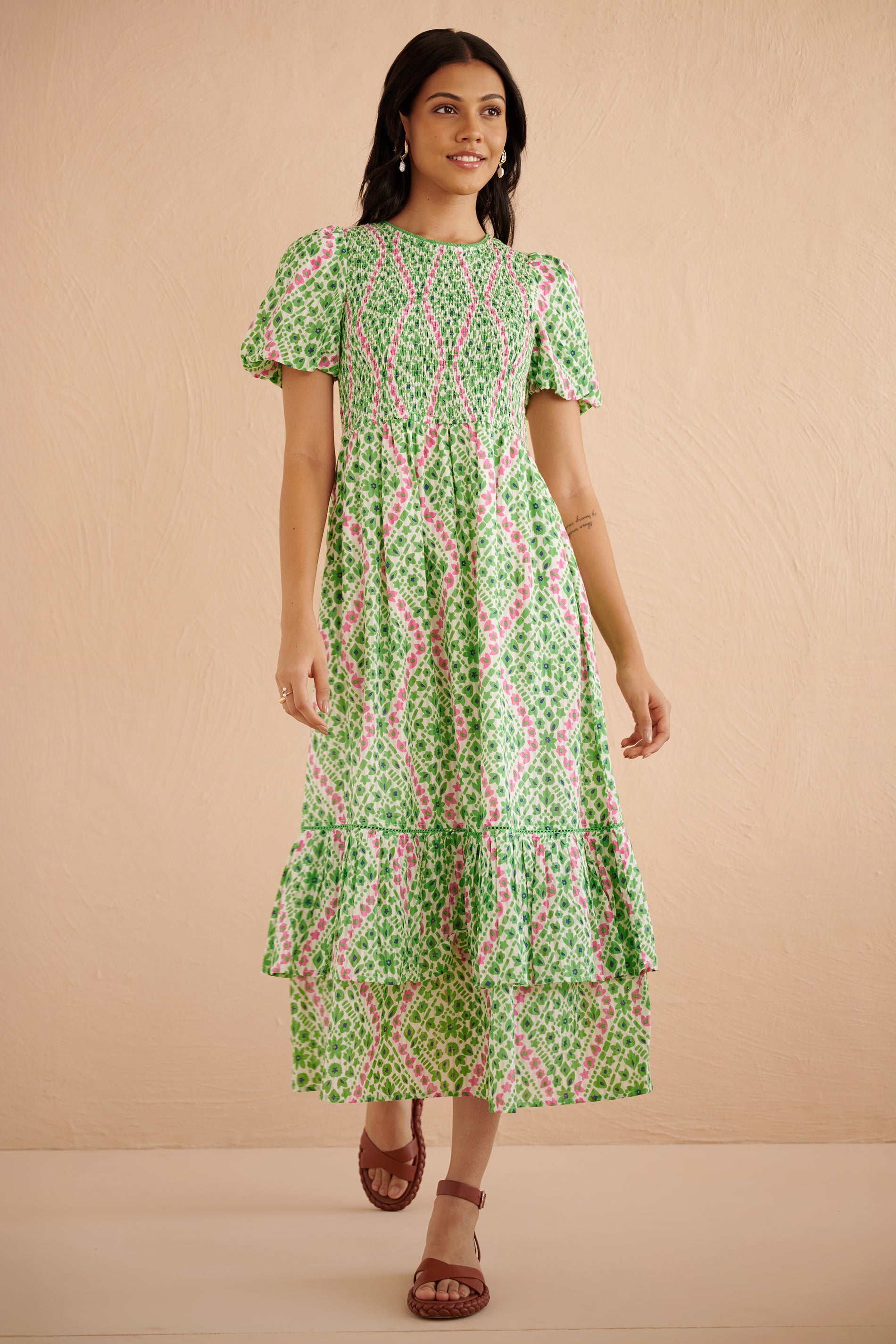 Front view of woman wearing the Quant midi dress in Athena Heart print, featuring a smocked bodice, puffed sleeves, tiered full skirt, lace trim, and side pockets in a green and pink floral-heart motif. Front view of woman wearing the Quant midi dress in Athena Heart print, featuring a smocked bodice, puffed sleeves, tiered full skirt, lace trim, and side pockets in a green and pink floral-heart motif.