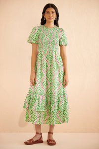 Front view of woman wearing the Quant midi dress in Athena Heart print, featuring a smocked bodice, puffed sleeves, tiered full skirt, lace trim, and side pockets in a green and pink floral-heart motif. Front view of woman wearing the Quant midi dress in Athena Heart print, featuring a smocked bodice, puffed sleeves, tiered full skirt, lace trim, and side pockets in a green and pink floral-heart motif.