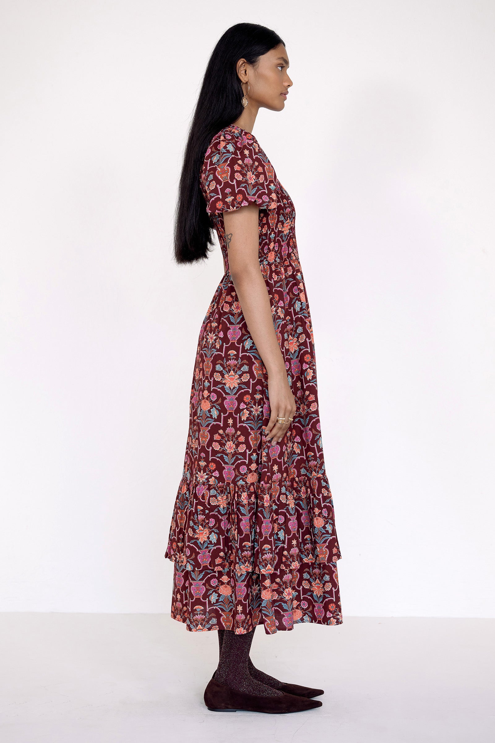 Side view of model standing. She is wearing the Quant dress in Floral Vase print. It features a smocked bodice, puffed sleeves, and a full tiered skirt with pockets.