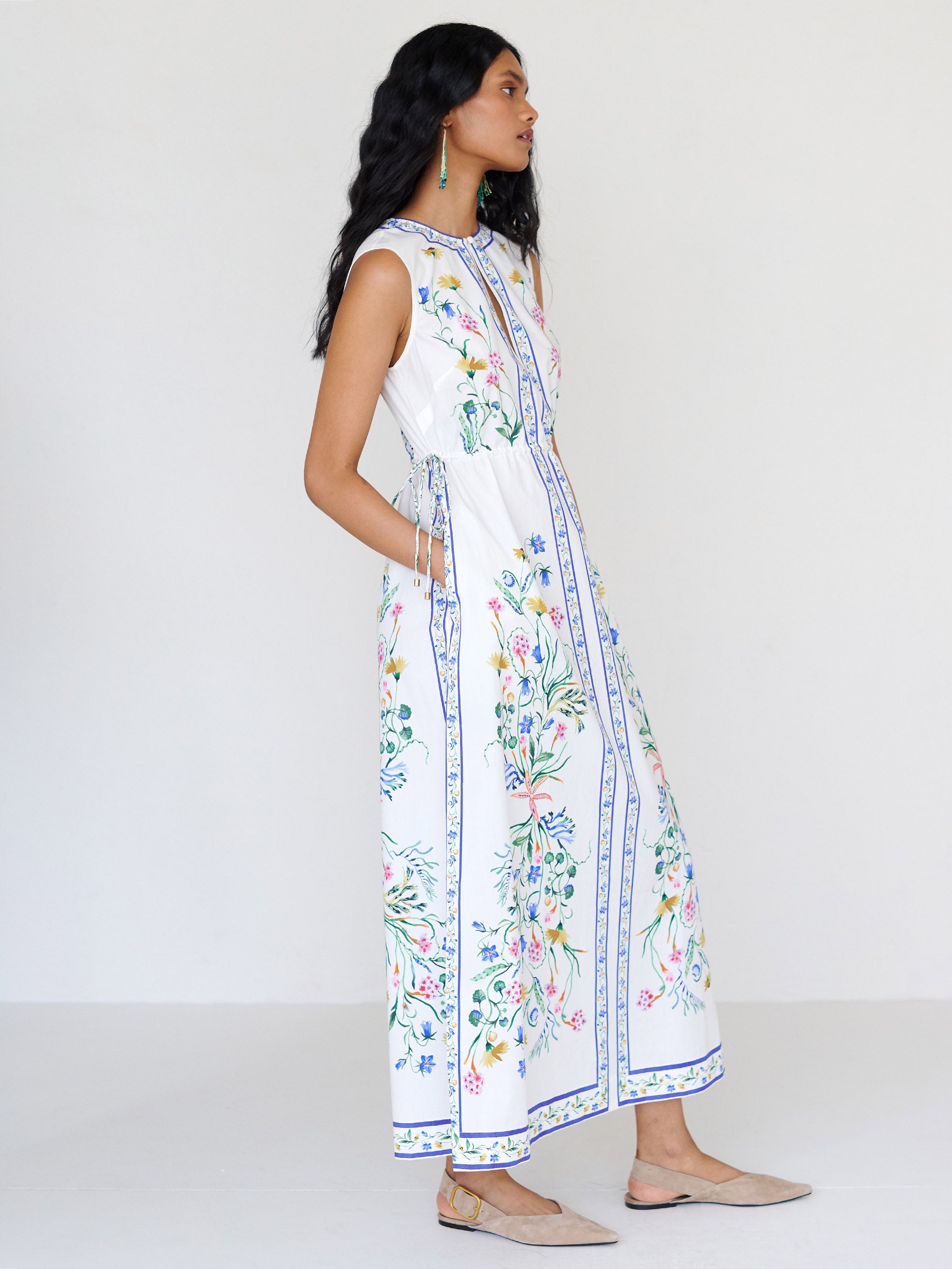 Side view close up of a woman standing with hands in pocket. She is wearing the Rhonda column dress in Tidepool print. It features a crewneck with a fabric-covered button and keyhole detail, side drawstrings with gold caps, and side-seam pockets.