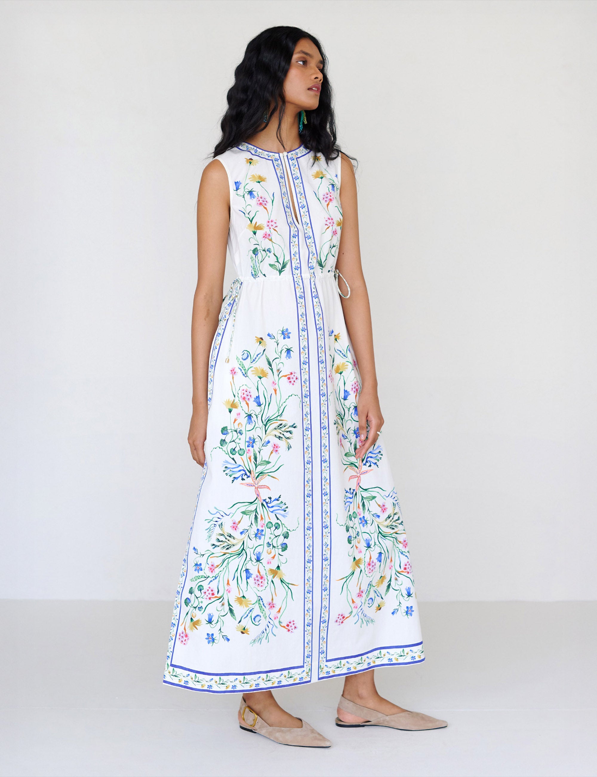 Front view of a woman standing looking sideways. She is wearing the Rhonda column dress in Tidepool print. It features a crewneck with a fabric-covered button and keyhole detail, side drawstrings with gold caps, and side-seam pockets.