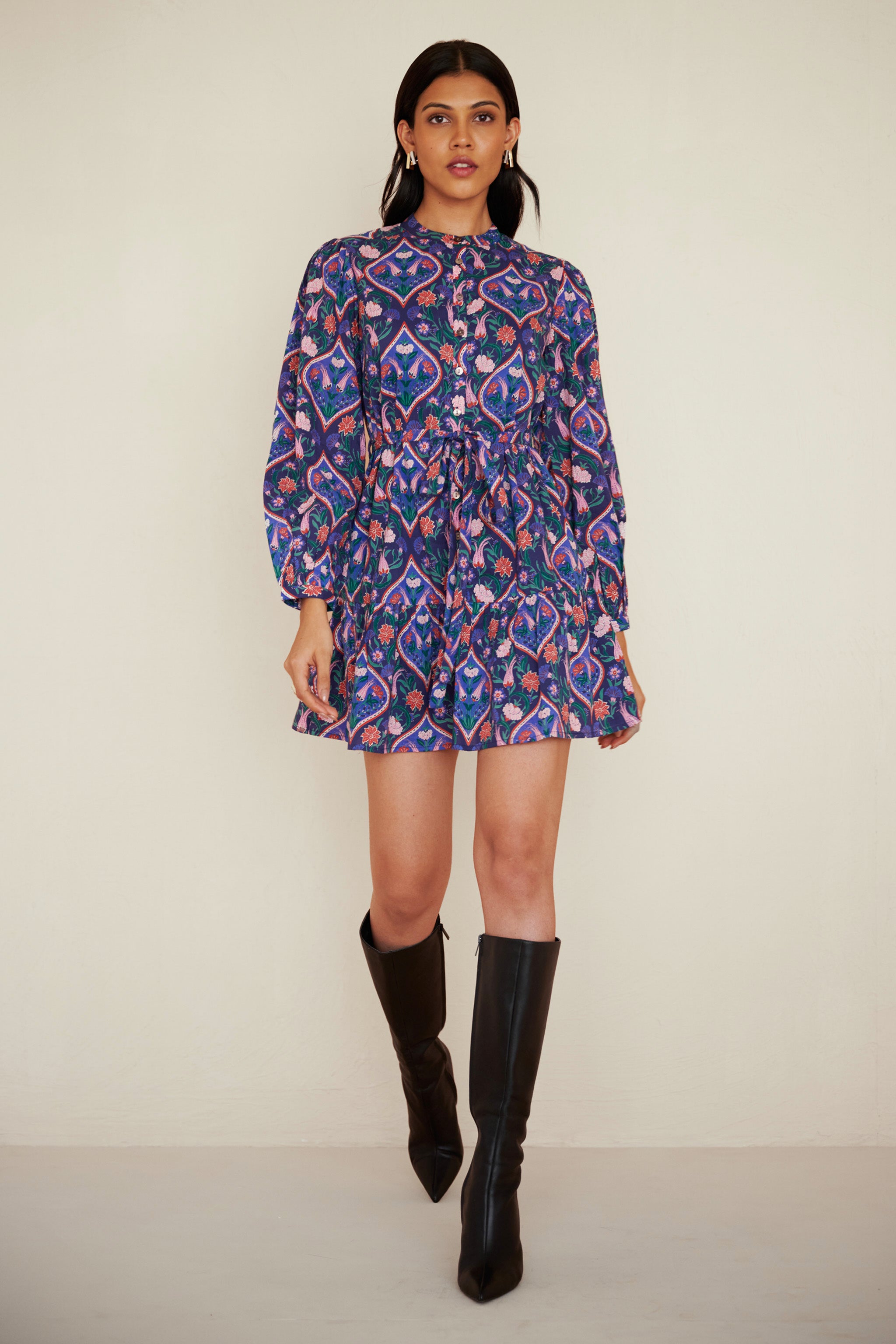 Front view of woman wearing a cotton mini dress with wrist-length sleeves, a floral tile print, and a detachable belt, styled with side pockets.