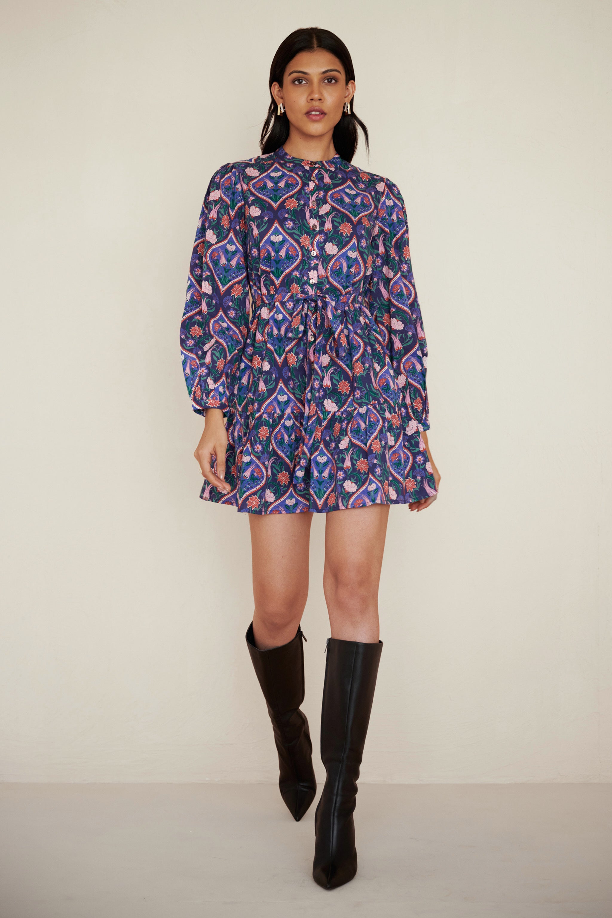 Front view of a woman walking. She is wearing Roma dress in Persian Tile Lapis print. Its a cotton mini dress with wrist-length sleeves, a floral tile print, and a detachable belt, styled with side pockets.
