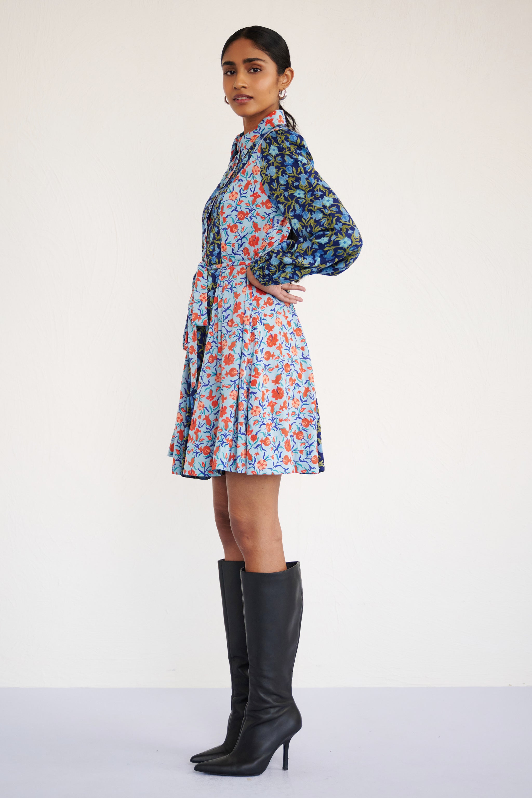 Side view of woman standing. She is wearing Rowena Dress in Pomegranate Vine print, Lapis color. It is a mini shirtdress with lantern sleeves, a delicate collar, and contrast pomegranate vine print, styled with side pockets.
