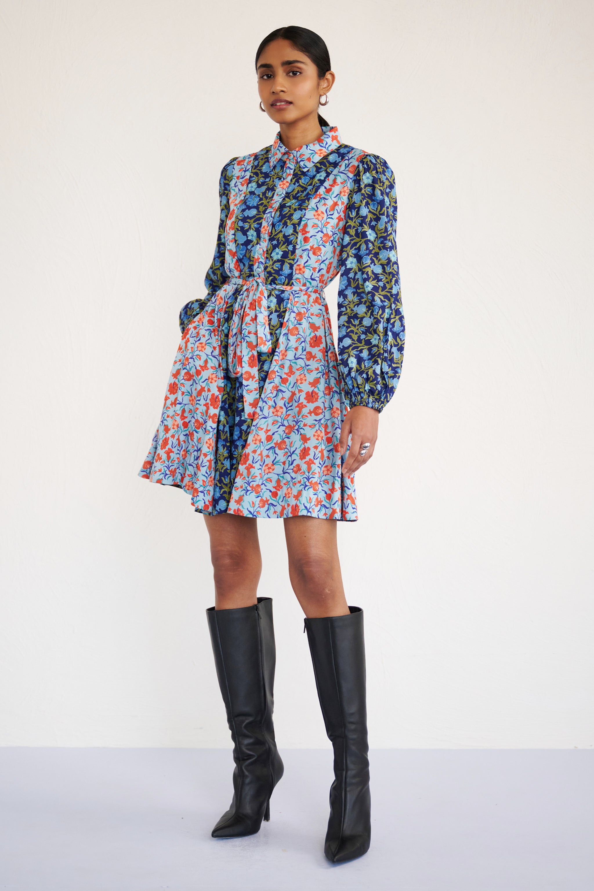 Front side view of woman standing. She is wearing Rowena Dress in Pomegranate Vine print, Lapis color with her left hand in pocket. It is a mini shirt dress with lantern sleeves, a delicate collar, and contrast pomegranate vine print, styled with side pockets.