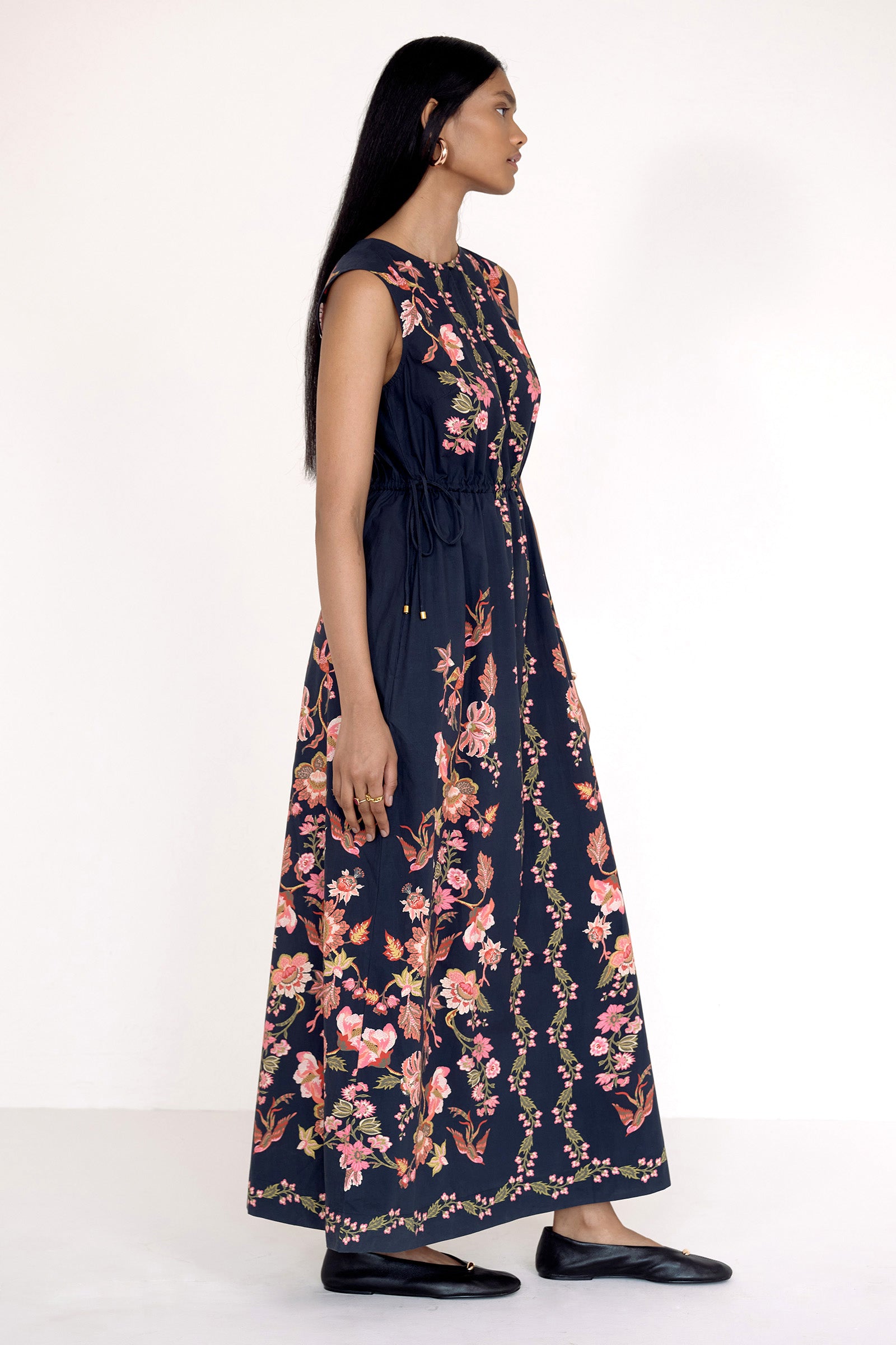 Side view of model standing. She is wearing the Rhonda dress in Chintz Jet. It features a crewneck with keyhole detail, adjustable drawstring sleeves, column silhouette, and vintage-inspired floral and bird print.