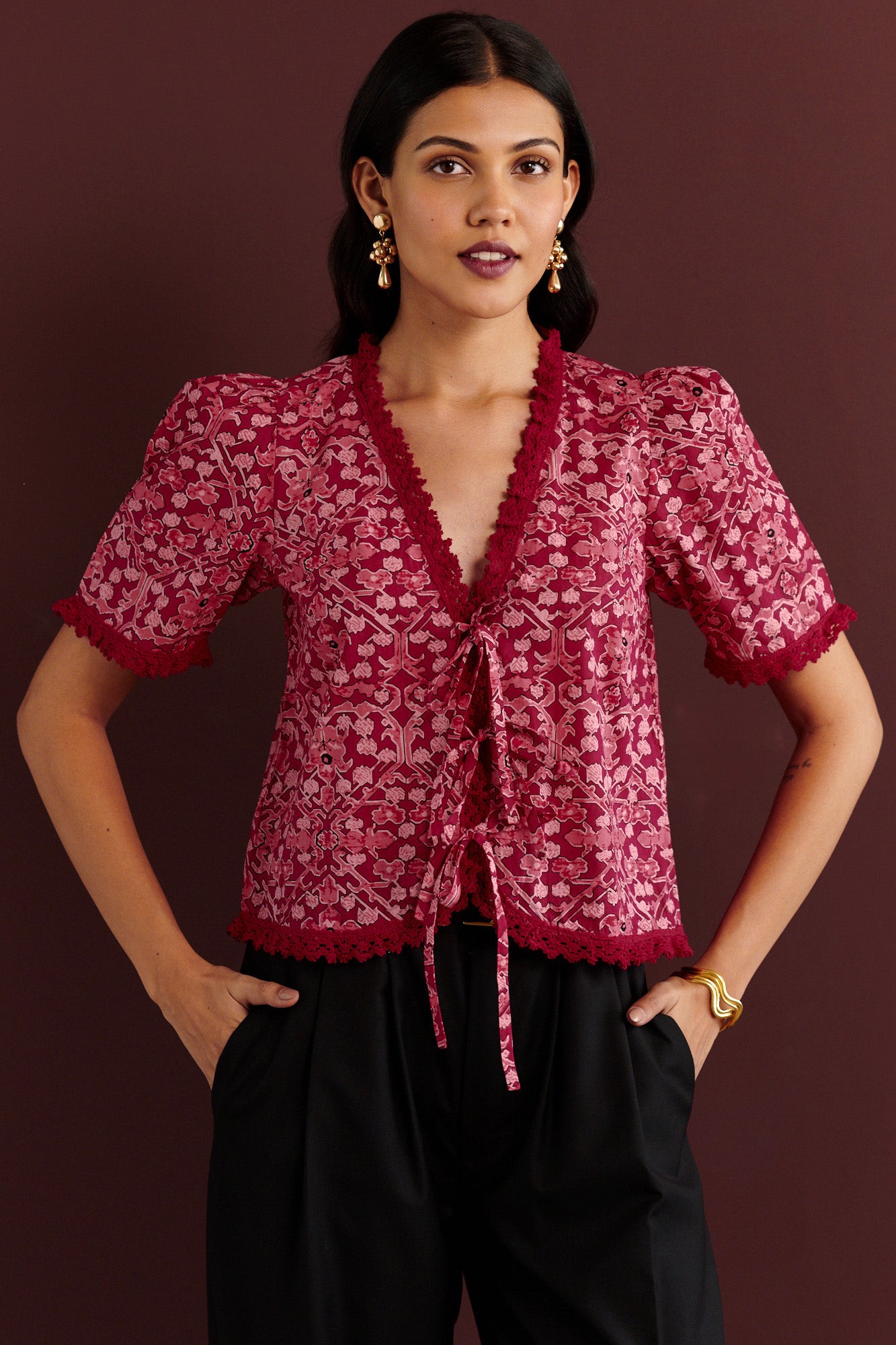 Front view of woman standing. She is wearing the Sarah top in the Ottoman print. It is a Berry red cotton cambric blouse with red lace trim and three front ties, styled for a festive and elegant winter look.