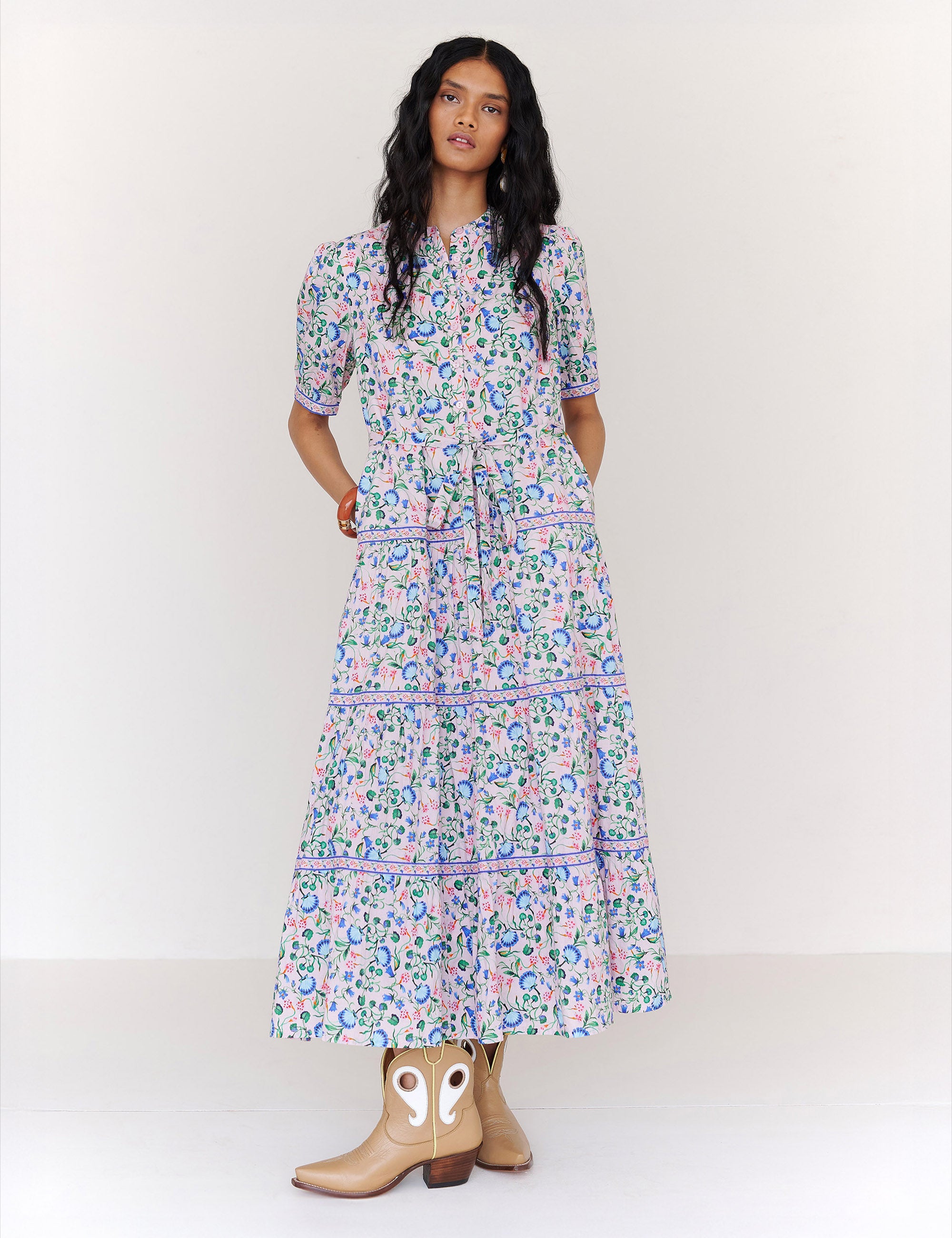 Front view of a woman standing. She is wearing the Saylor dress in Clam Garland floral print with her hands in pocket. It features a classic shirtdress design with stand collar, tiered skirt, contrast trim, and a detachable belt.