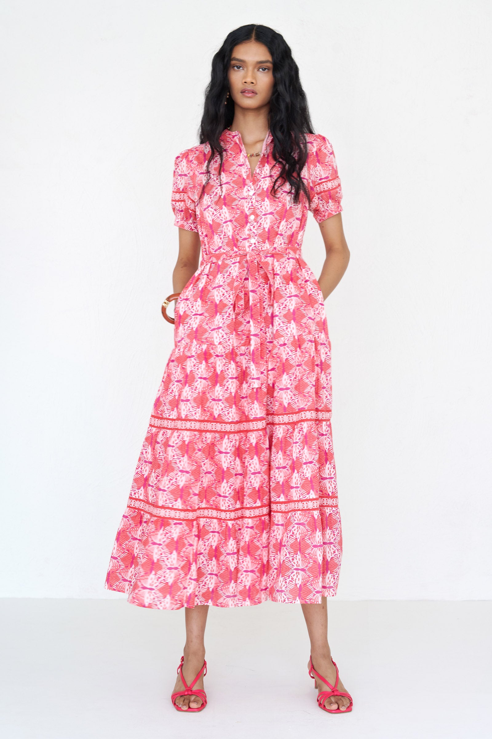 Front view of a woman standing wearing the Saylor dress in Flutter print with her hands in pocket. It features a stand collar, tiered skirt, detachable belt, and celtic-knot ladder lace detailing in a full-length shirtdress silhouette.